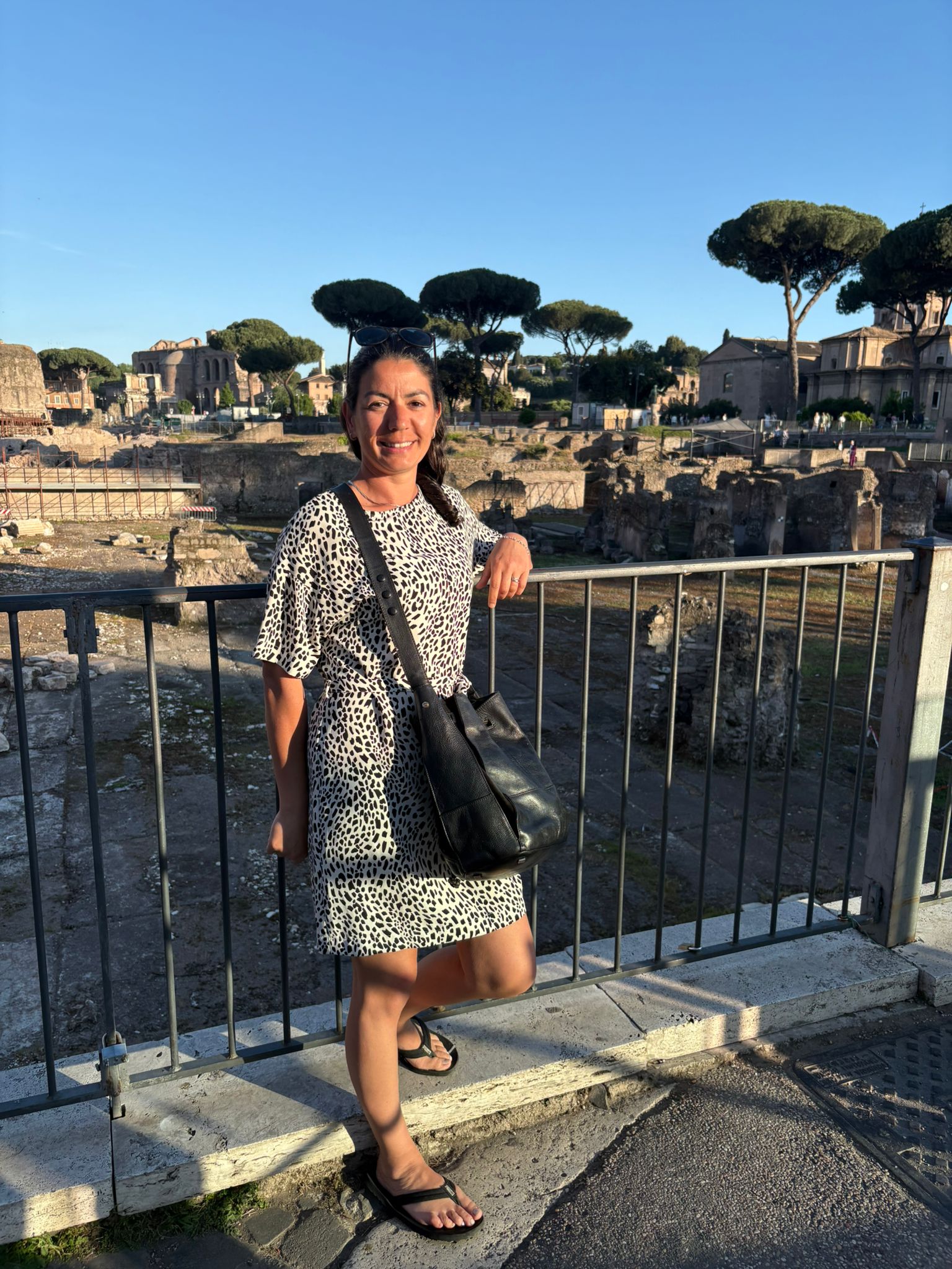 Person in patterned dress by ancient ruins and trees, leaning on fence with a black bag.