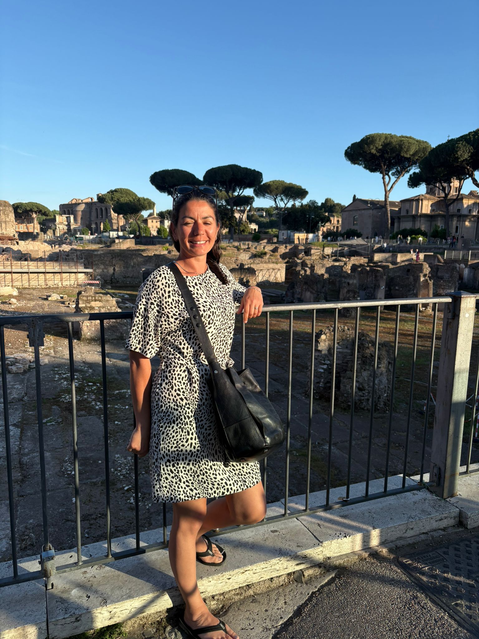 Person in patterned dress and sandals by railing, smiling at ancient ruins under blue sky.