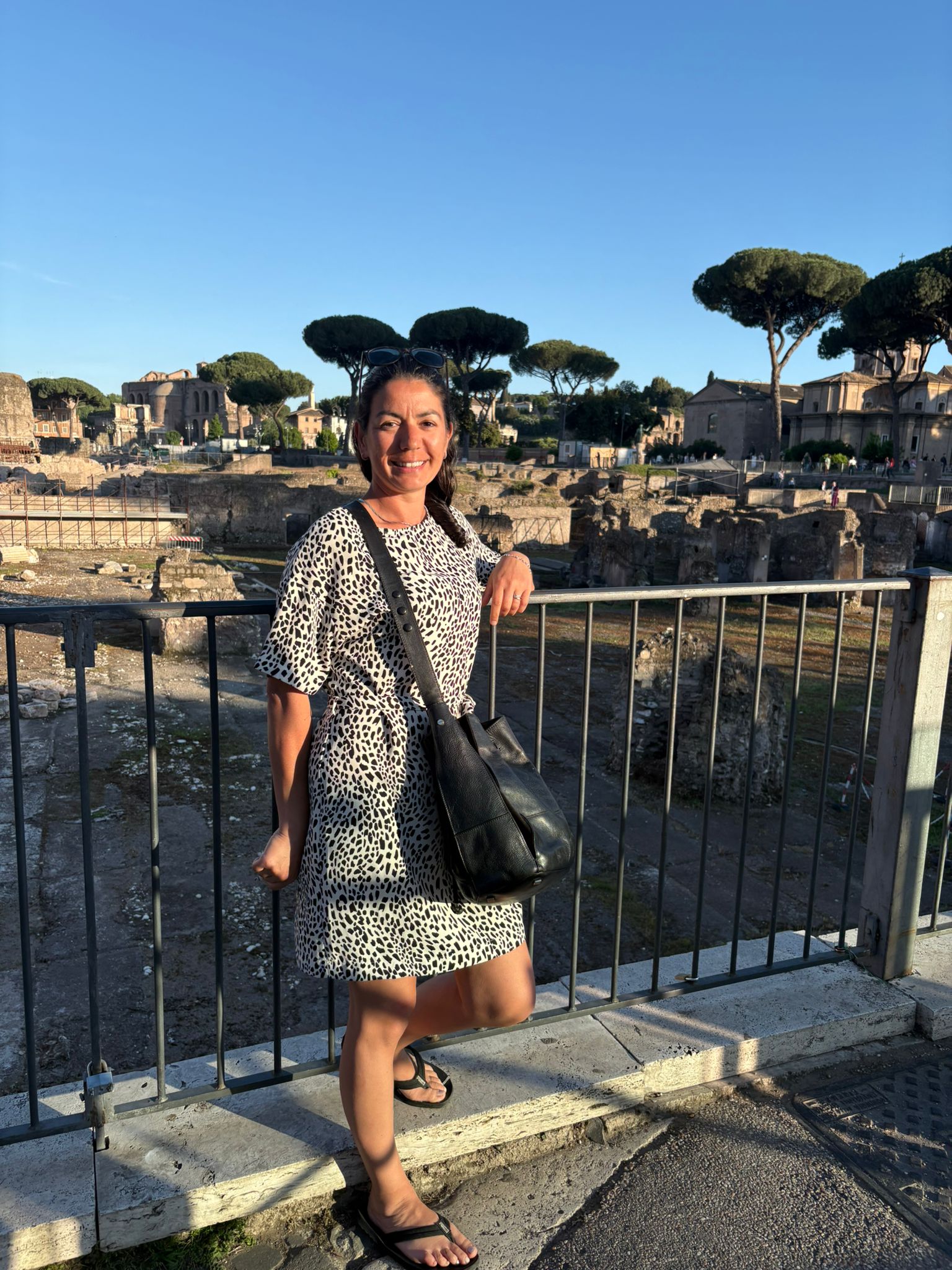 Person in patterned dress by railing, ancient ruins, trees, buildings, clear sky background.