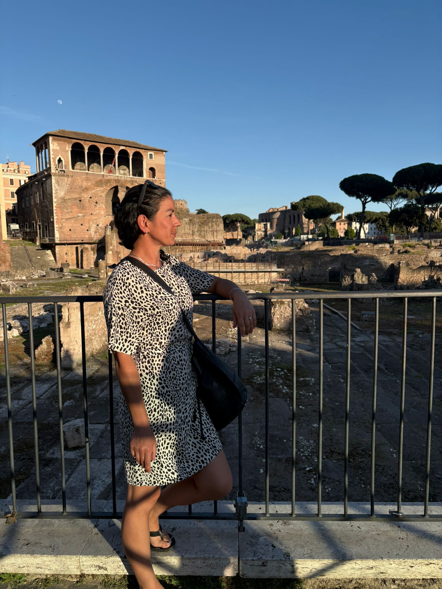 Person in patterned dress by railing with ruins, brick building, trees, clear sky in background.