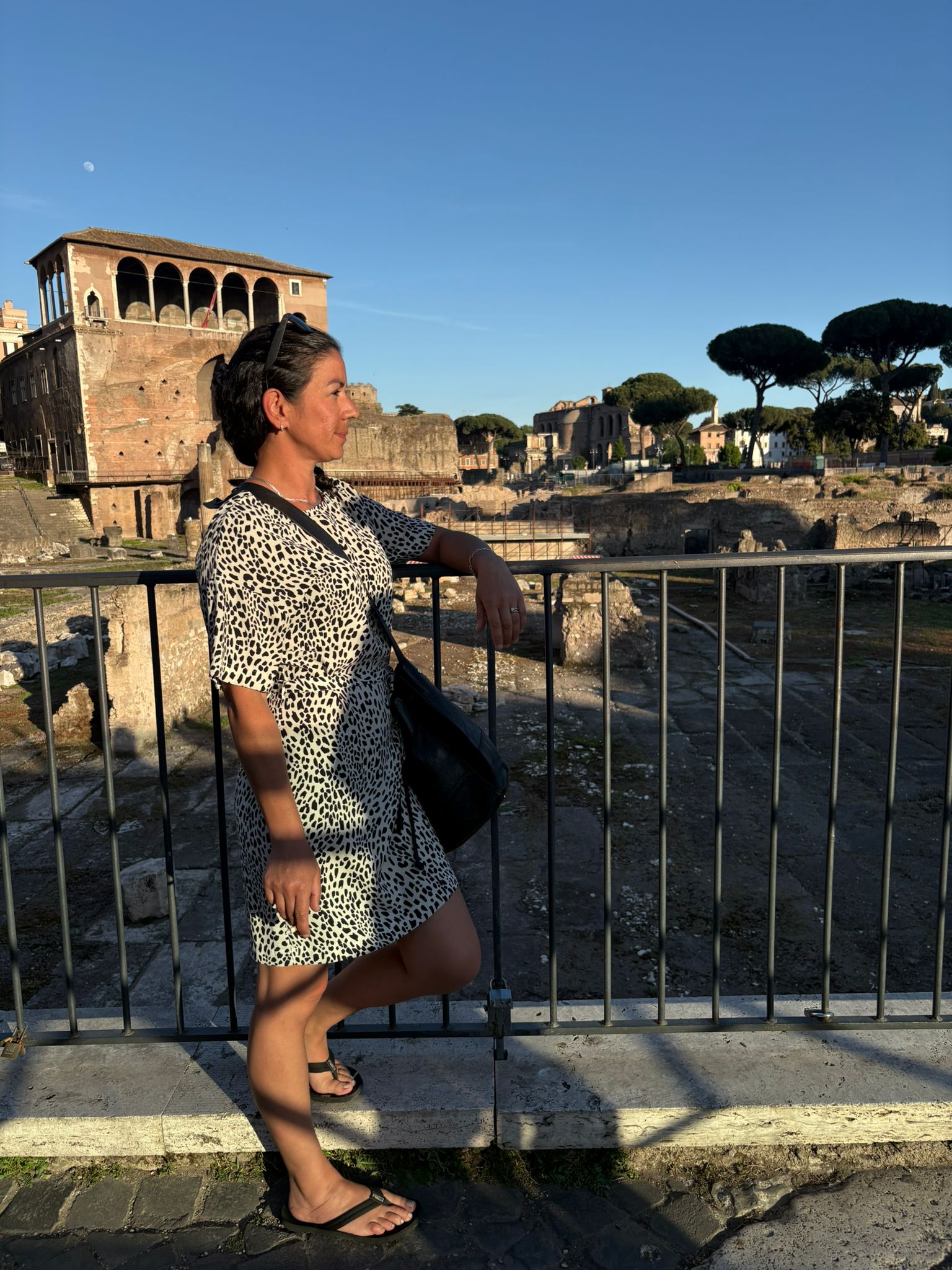 Person in a patterned dress by railing, overlooking ancient ruins and trees under clear sky.