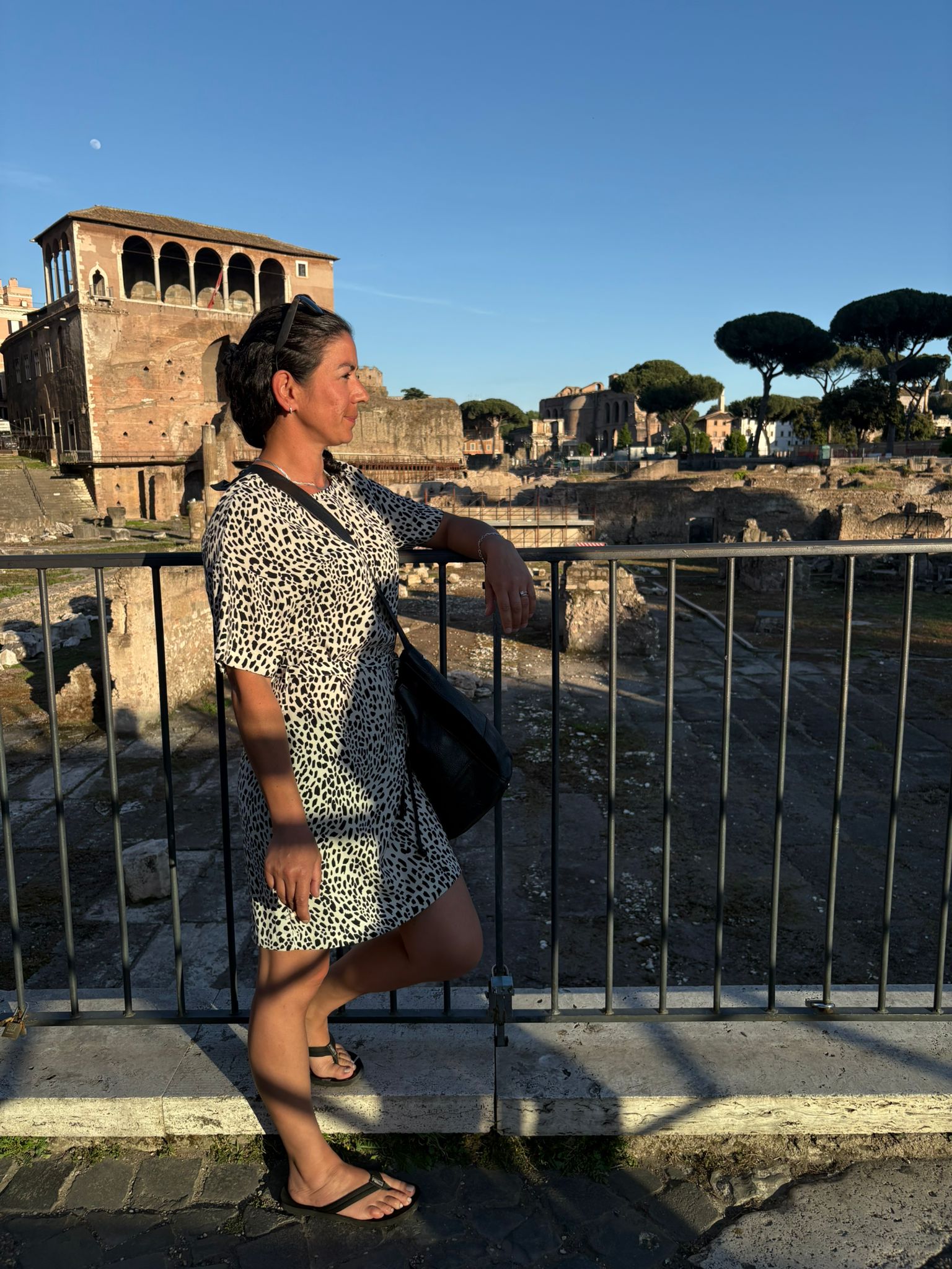 Person in patterned dress and sandals by railing, overlooking ancient ruins with a clear sky.