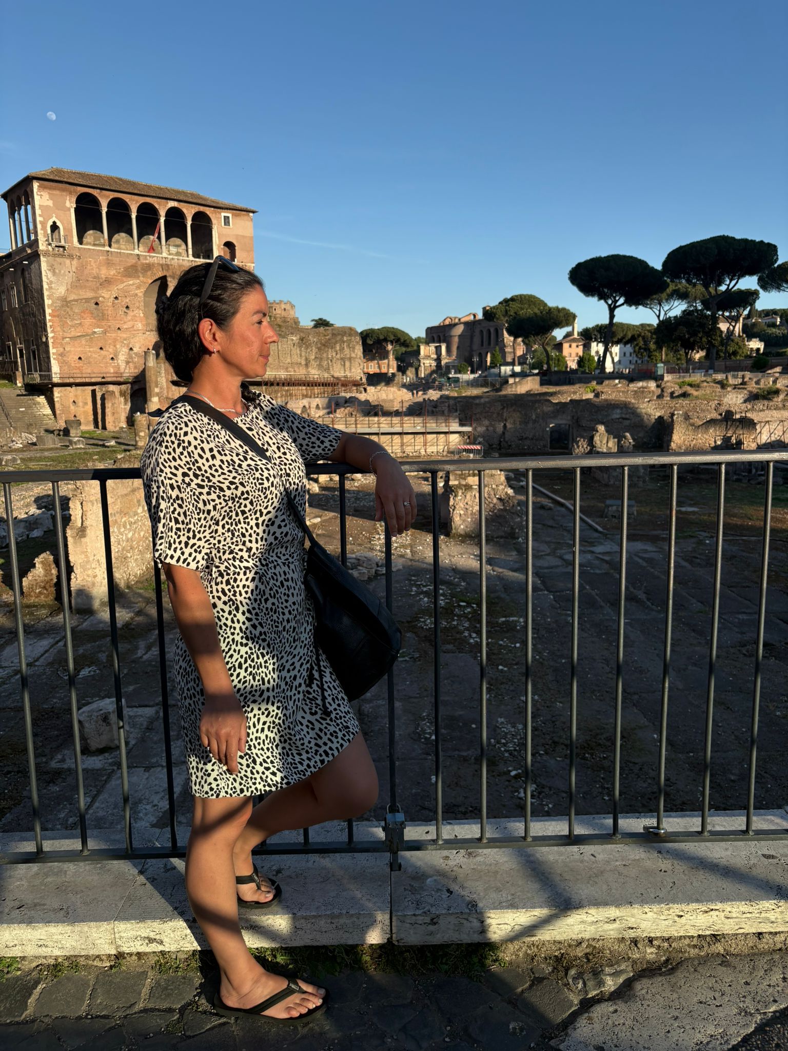 Person in patterned dress, by railing, overlooking ancient ruins and trees under blue sky.