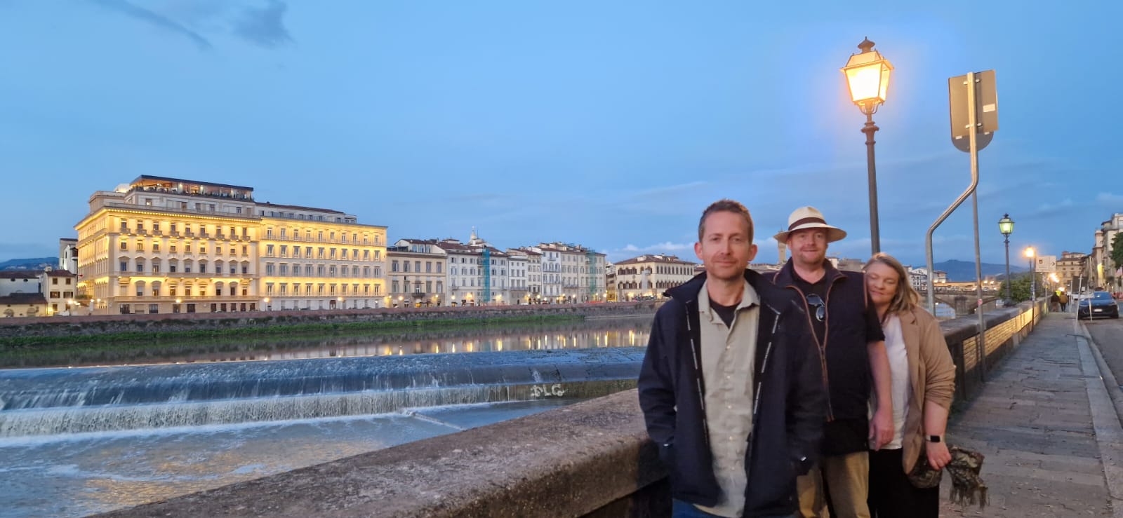 Three people on riverside promenade, lit building, flowing river, evening sky, streetlights.