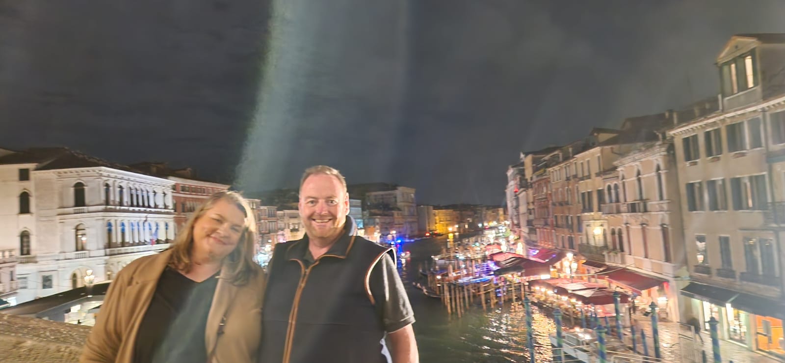 Man and woman on a bridge, canal and illuminated buildings in the background at night.