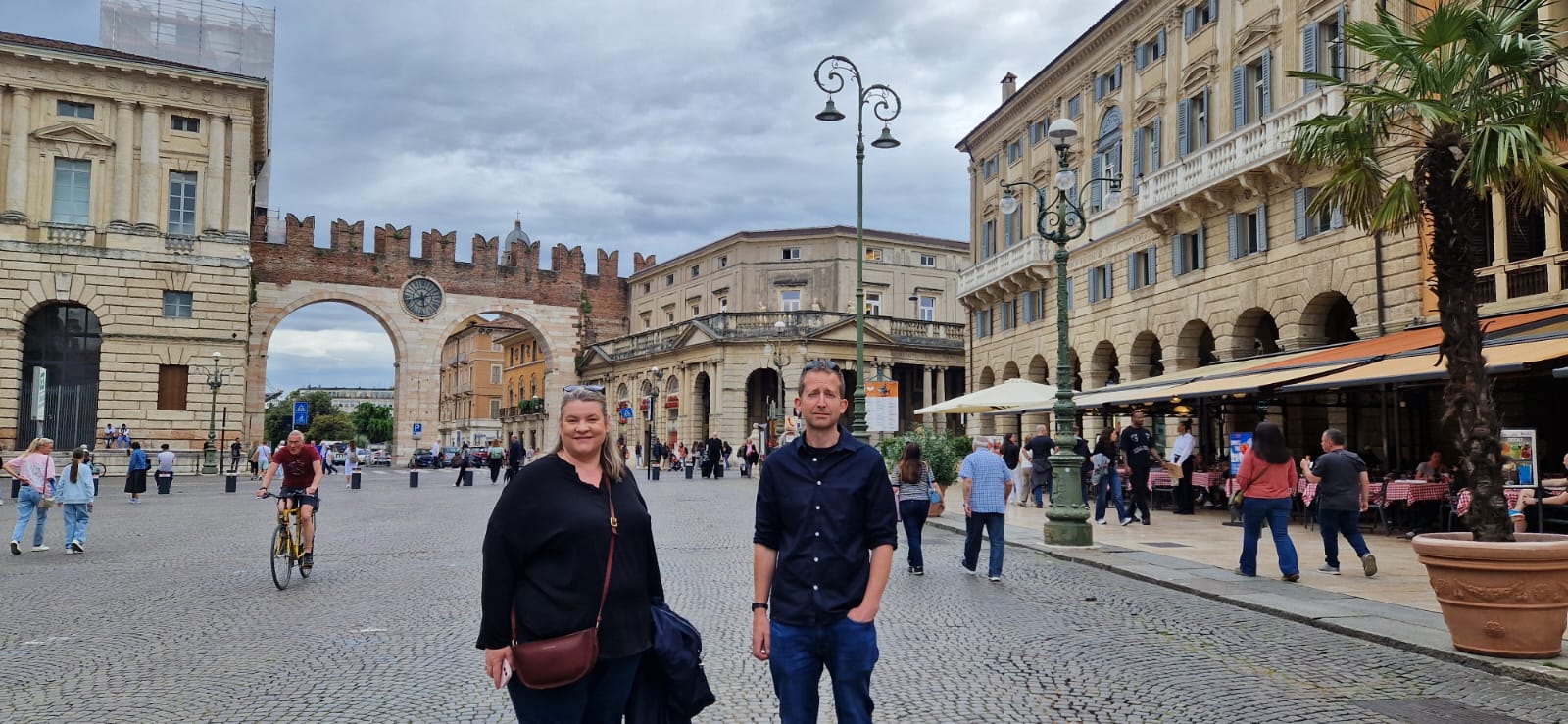 Historic European Piazza Scene European piazza with people, historical archway, cobblestones, cloudy sky, and café seating.