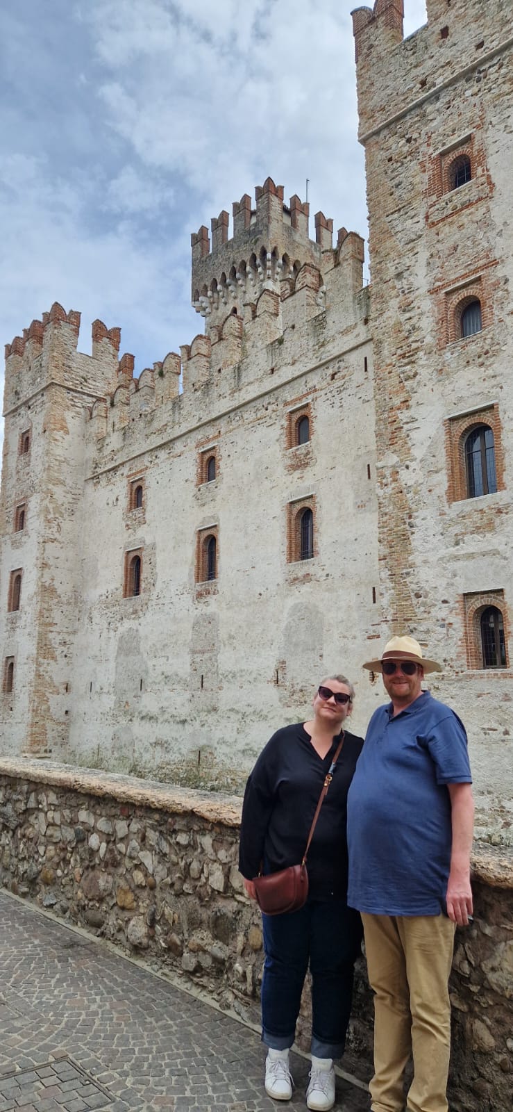 Two people smiling on walkway in front of a large stone castle with towers and cloudy sky.