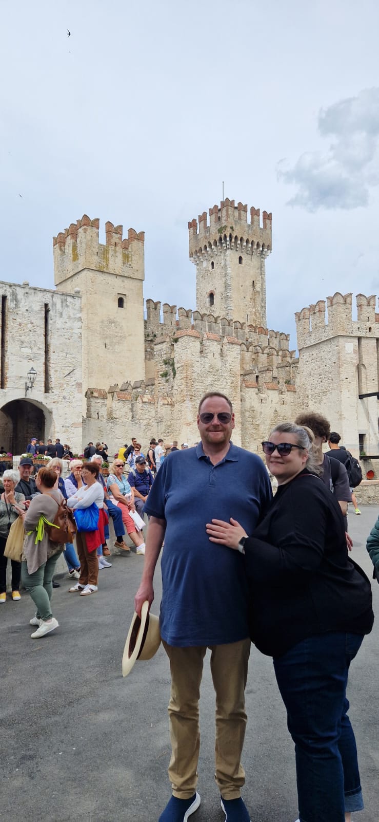 A couple in sunglasses stands by an ancient stone castle with tourists and overcast sky.