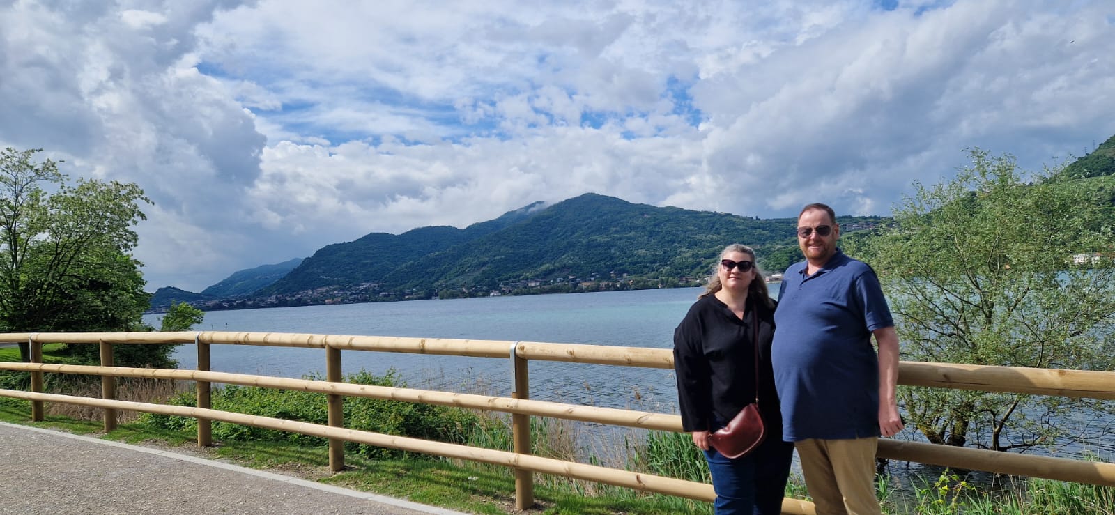 Couple on lakeside path with railing, lush hills, and cloudy sky in the background.