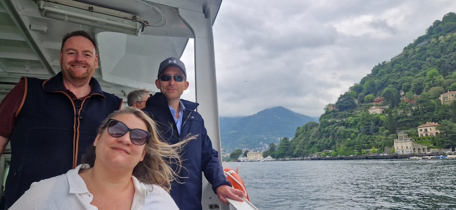 Group on a boat with hillside homes and greenery in the background under an overcast sky.