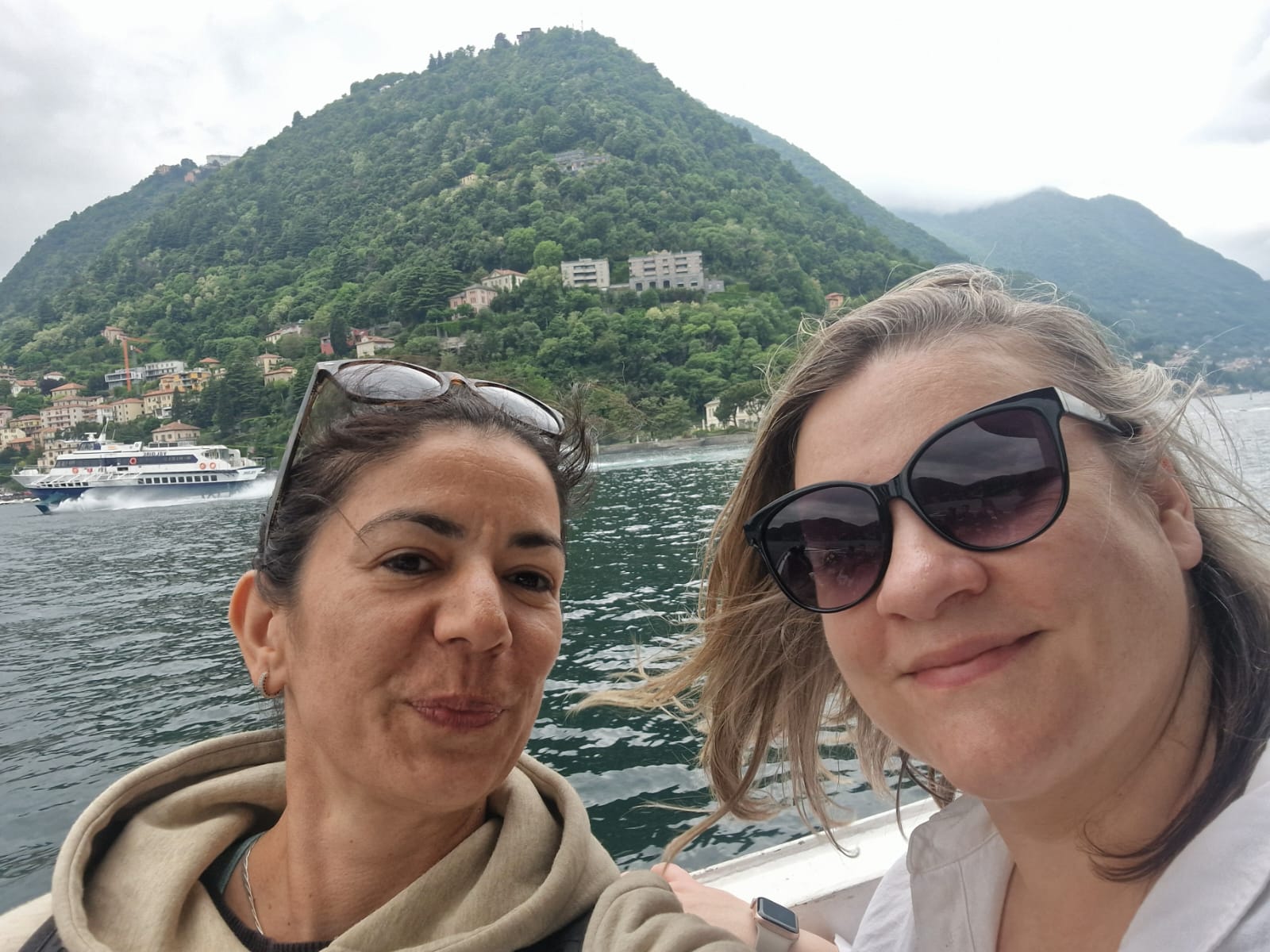 Two people taking a selfie on a boat, with hillside, buildings, and a ferry in the background.