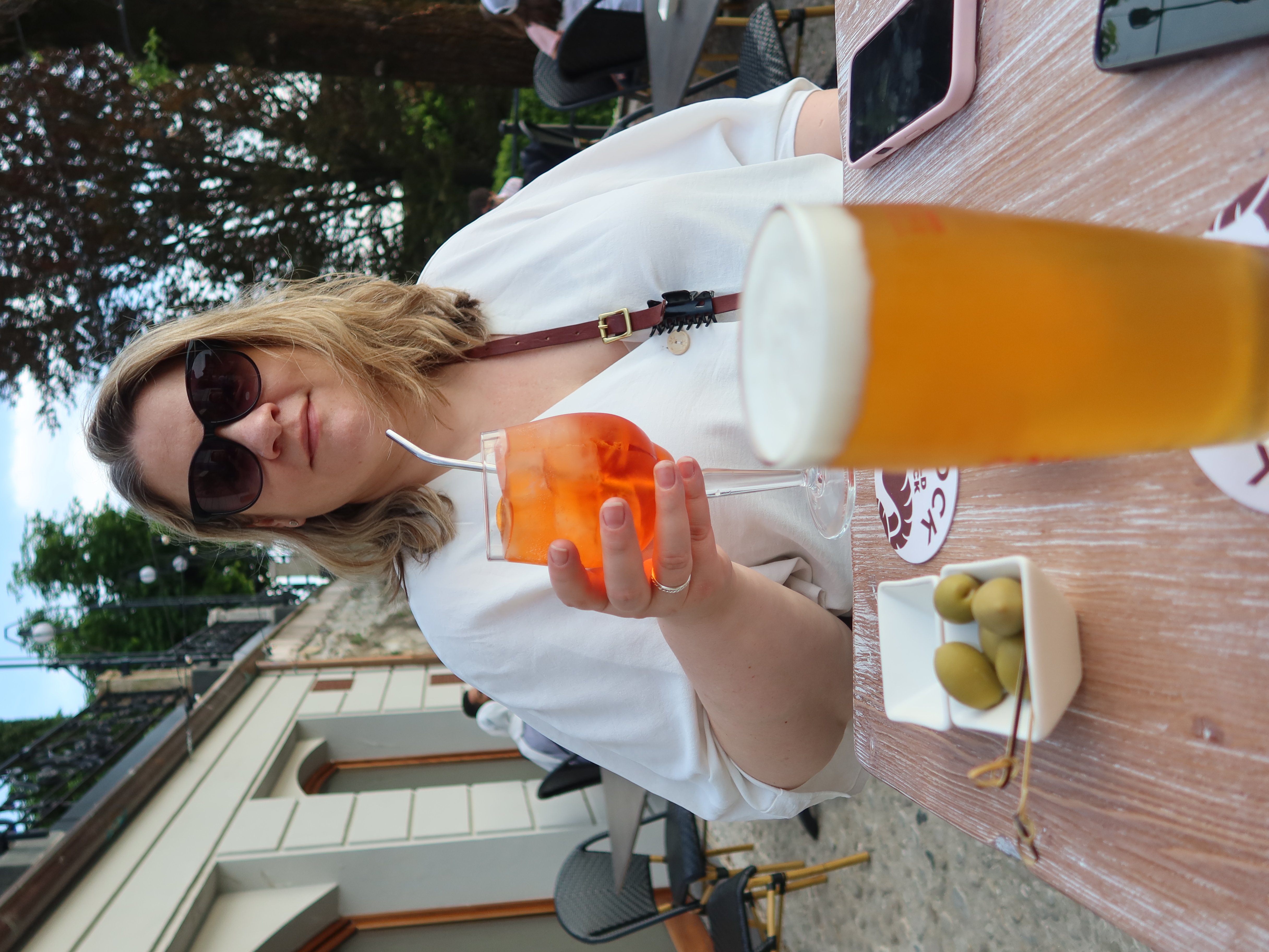 Person at café with orange drink; table has beer, olives, smartphone; building and trees behind.