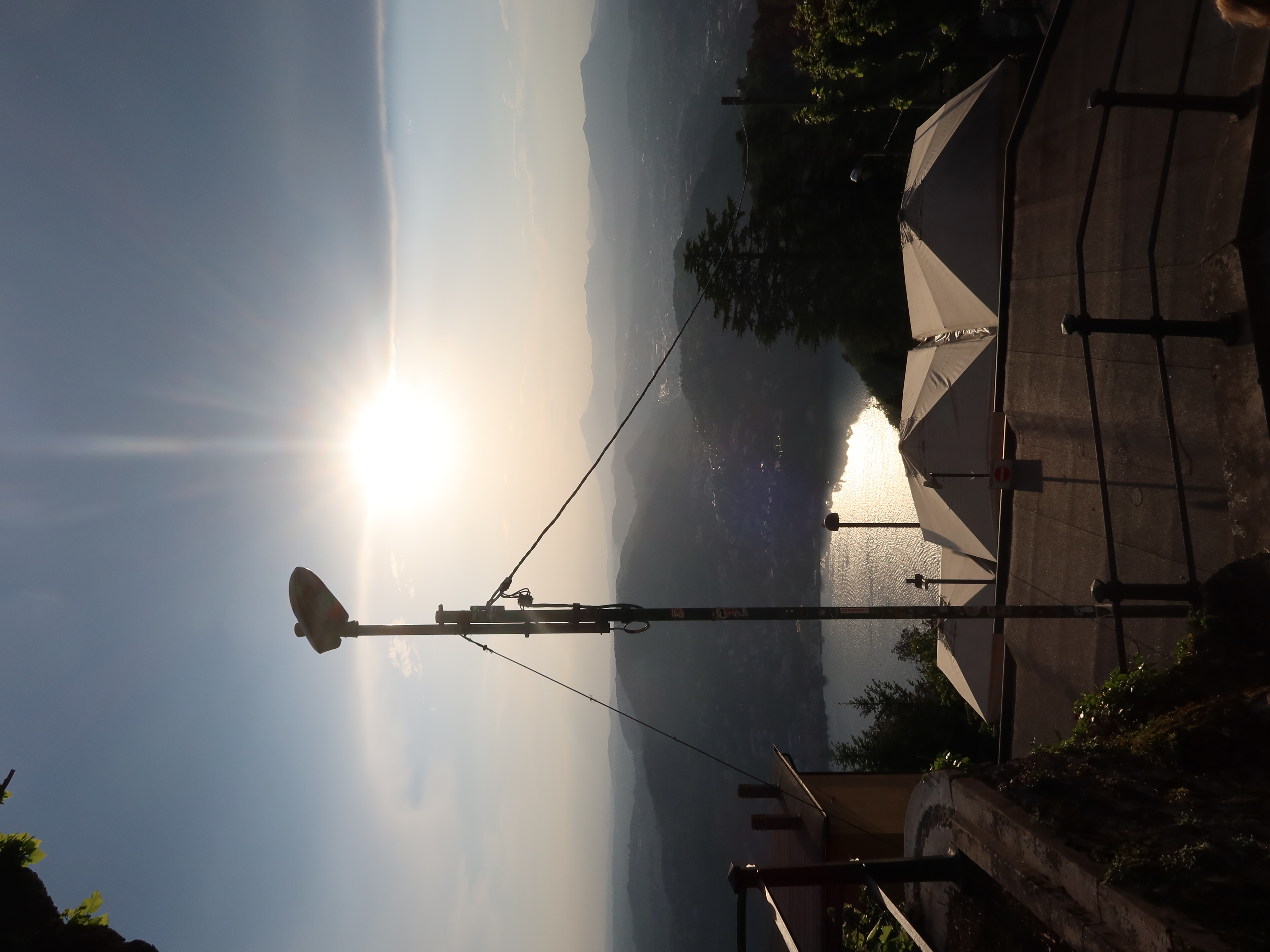 Streetlamp on hillside terrace overlooking water, tents below, sun shining, cloudy sky, mountains.