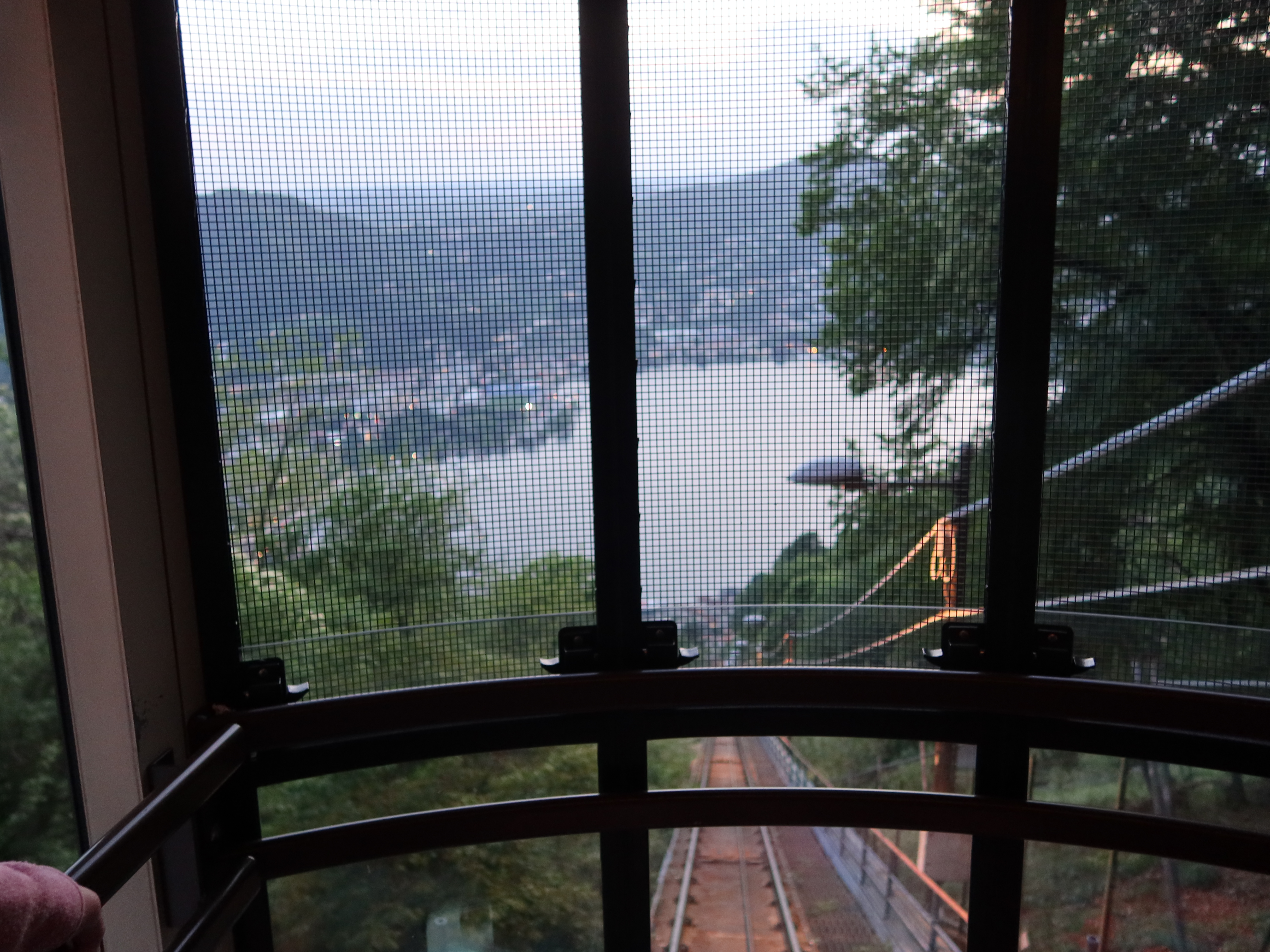 View from inside a funicular car through mesh, showing tracks, trees, and a lake with hills.