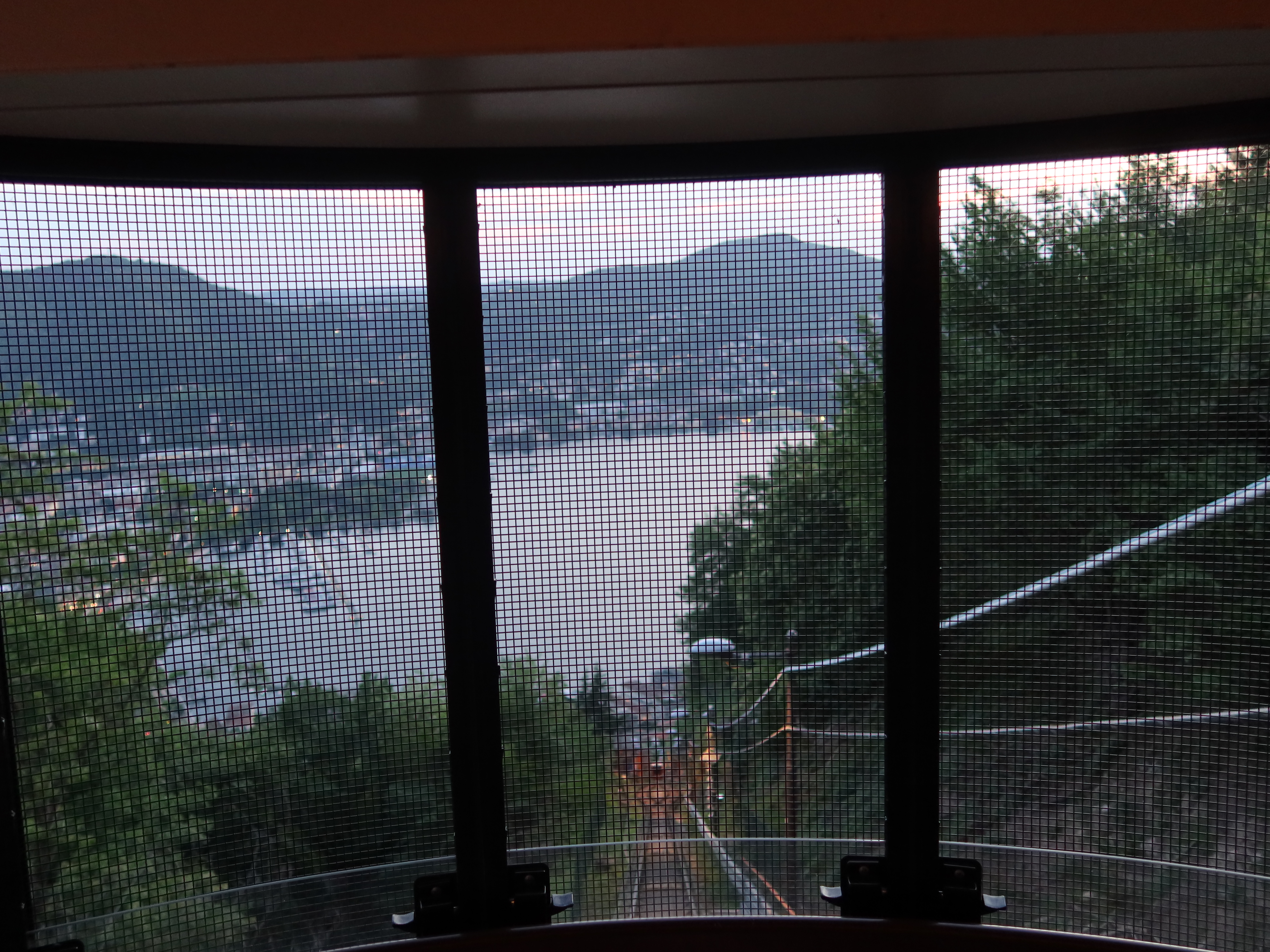 View from funicular cabin through gridded window, showing track, trees, hills, and water.