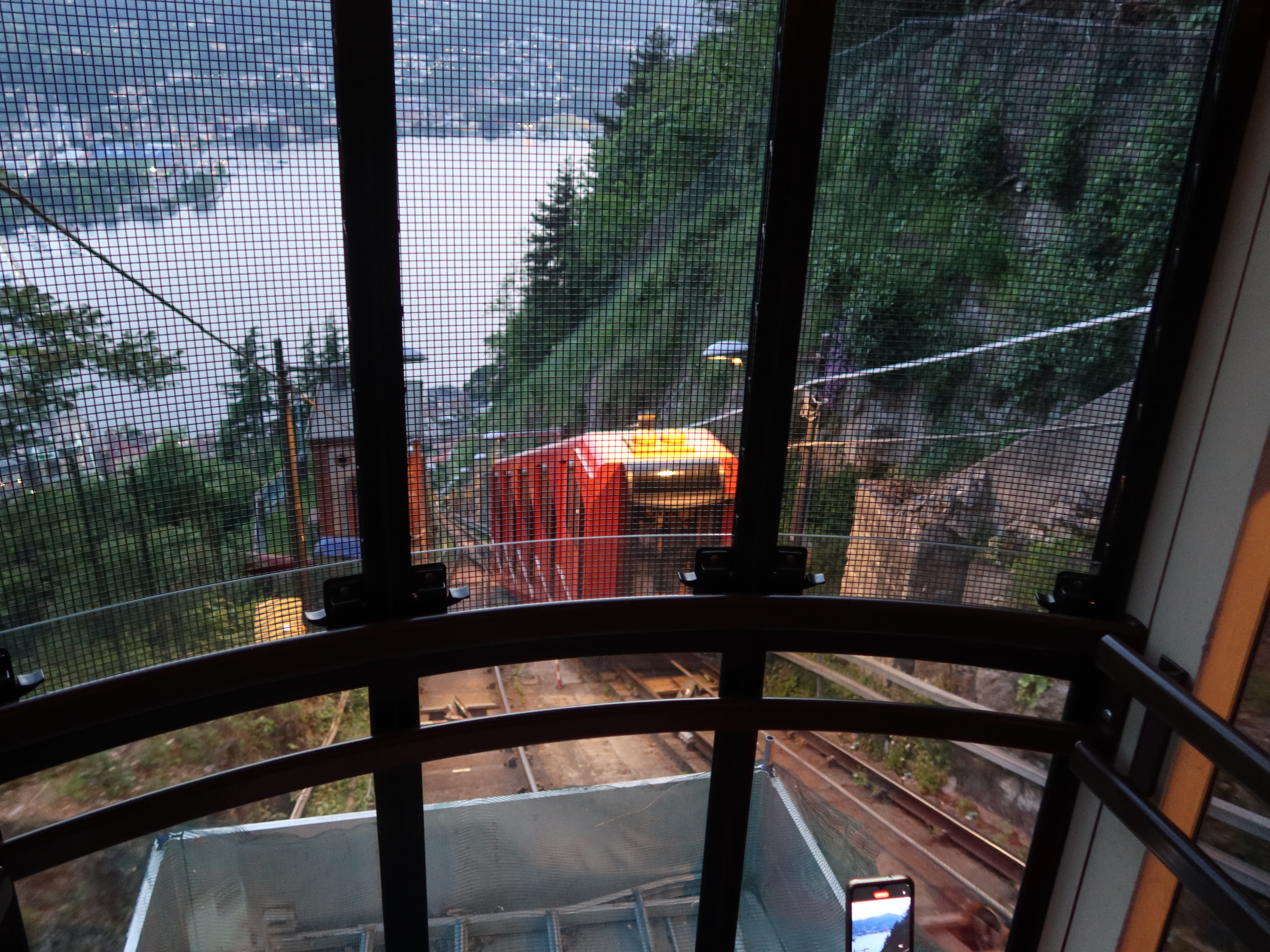 Hillside Funicular Overlooking Scenic Lake Red funicular climbs hillside, greenery surrounds; view over water, distant buildings, phone foreground.