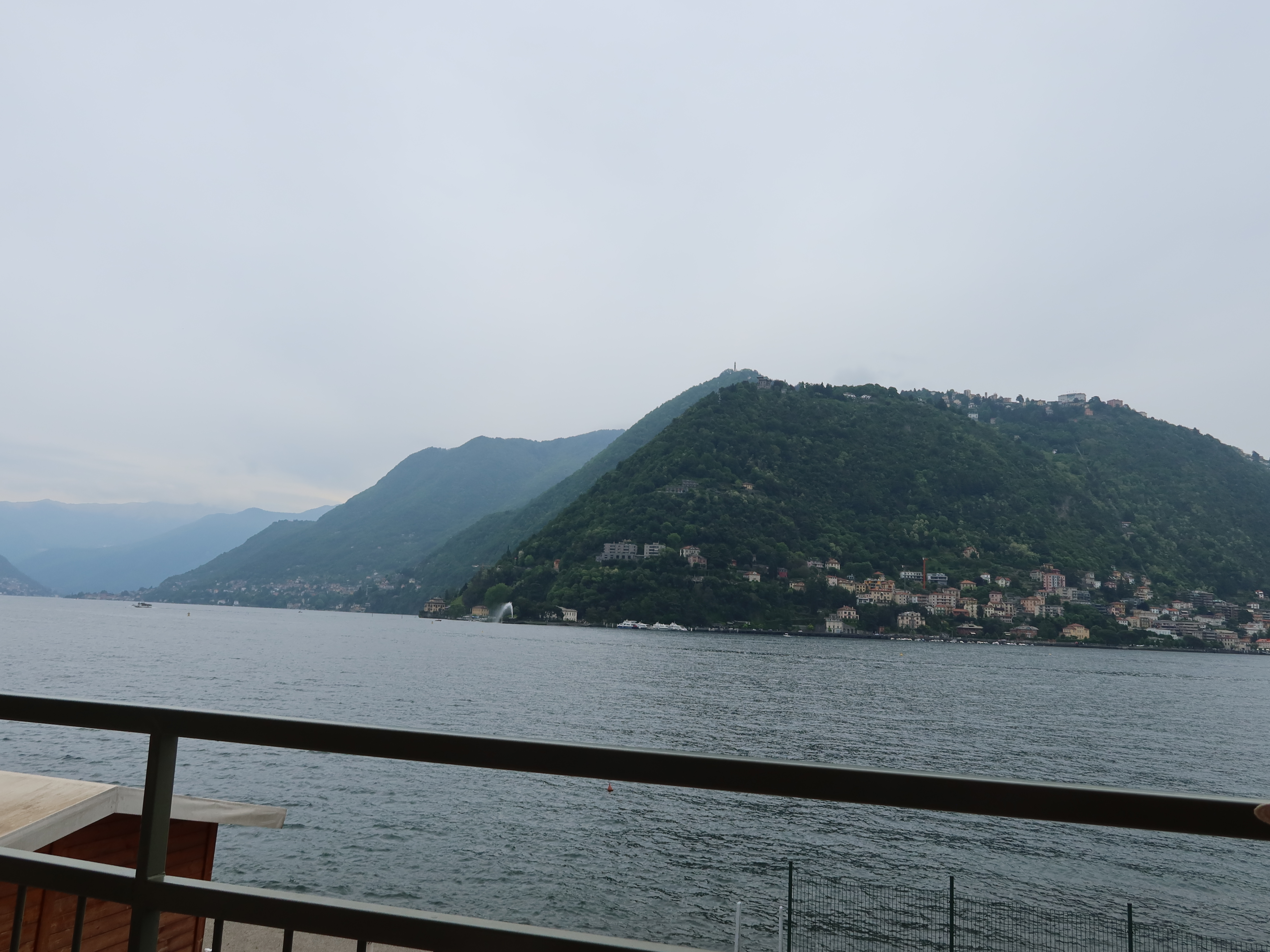 Large lake with railing in foreground, forested hills, scattered buildings, overcast sky.