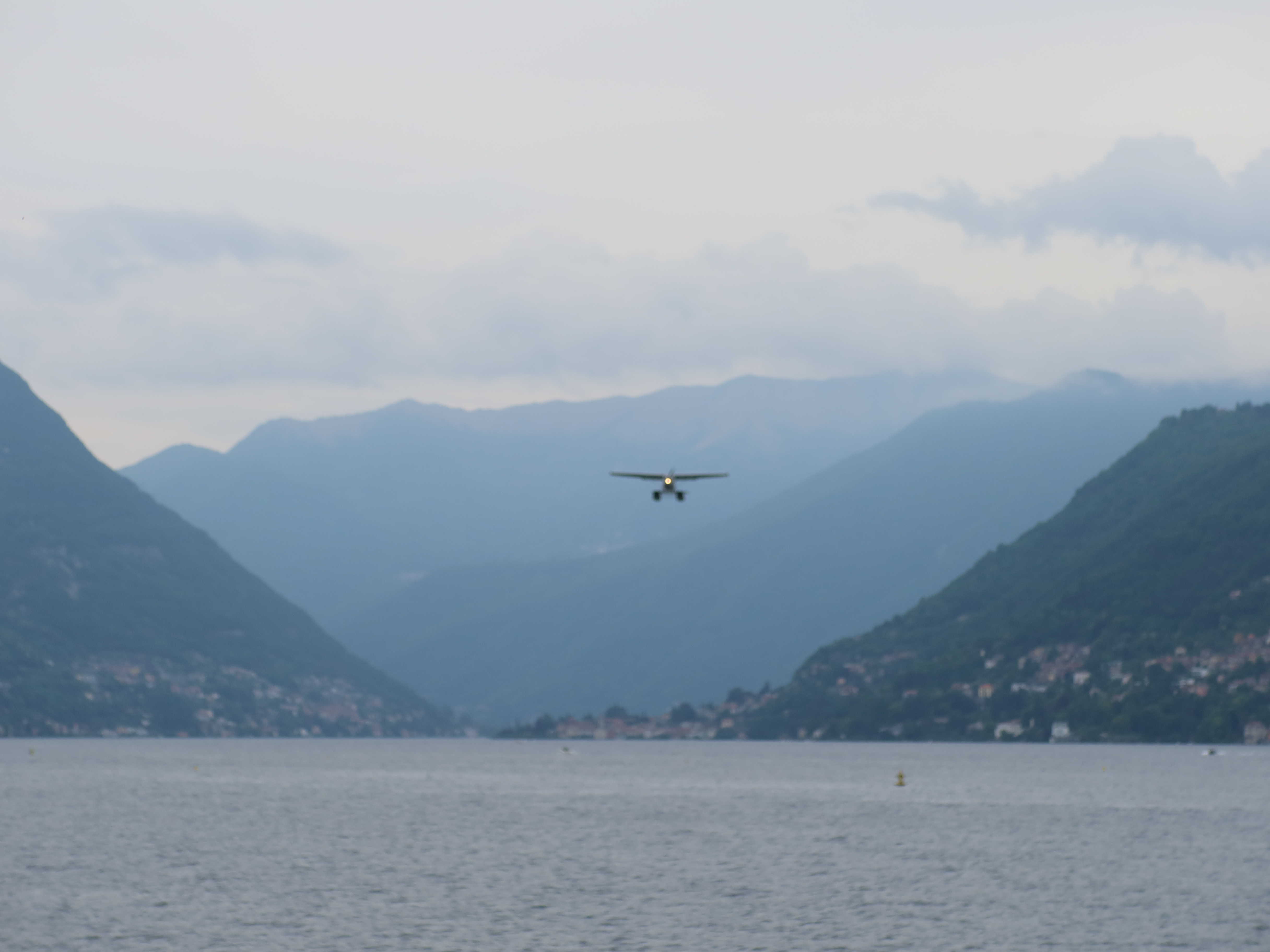 Small aircraft flying over water near green hills and distant mountains under a cloudy sky.