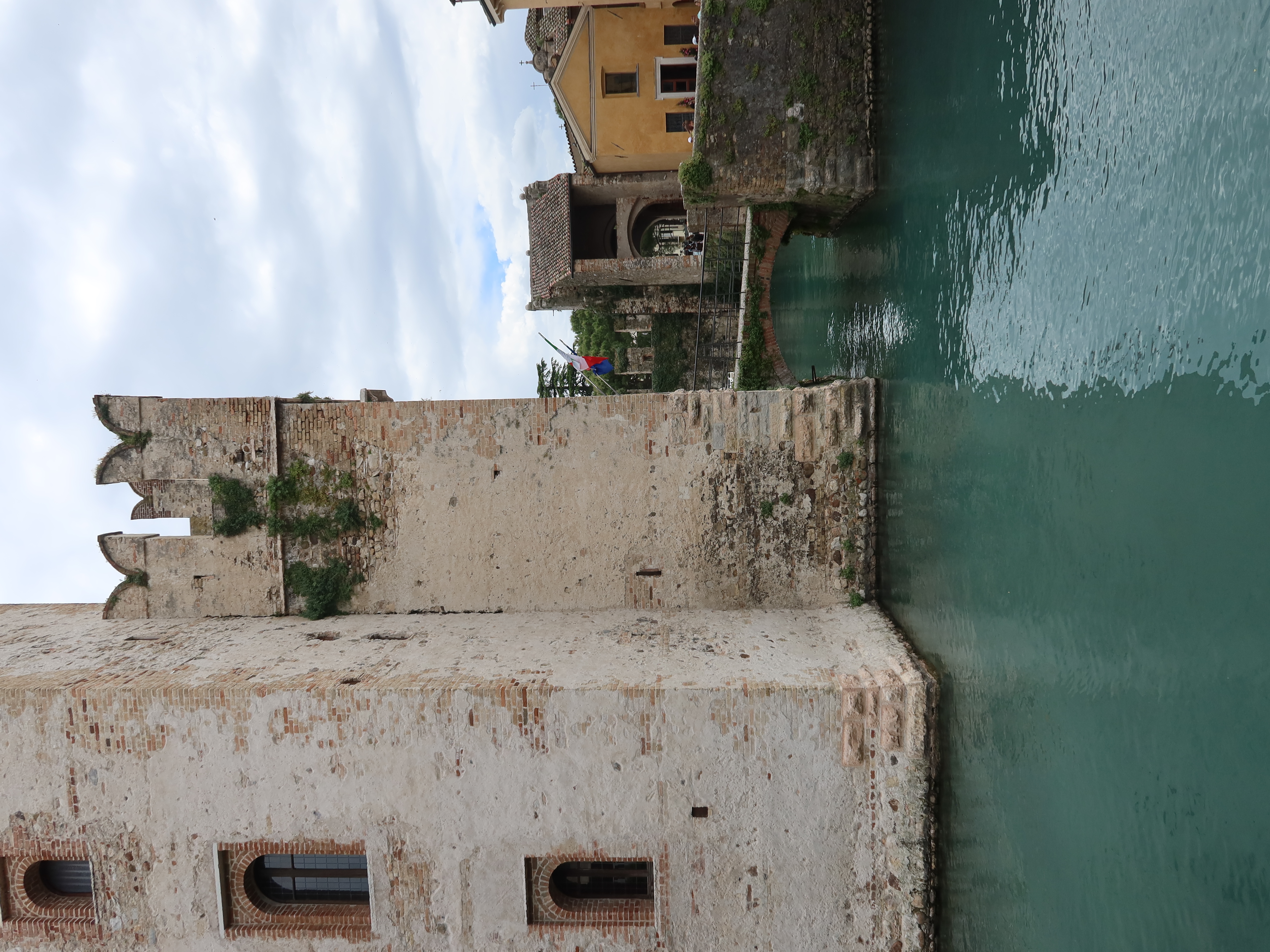 Historic stone building with crenellations by turquoise water, adjacent to a yellow structure.