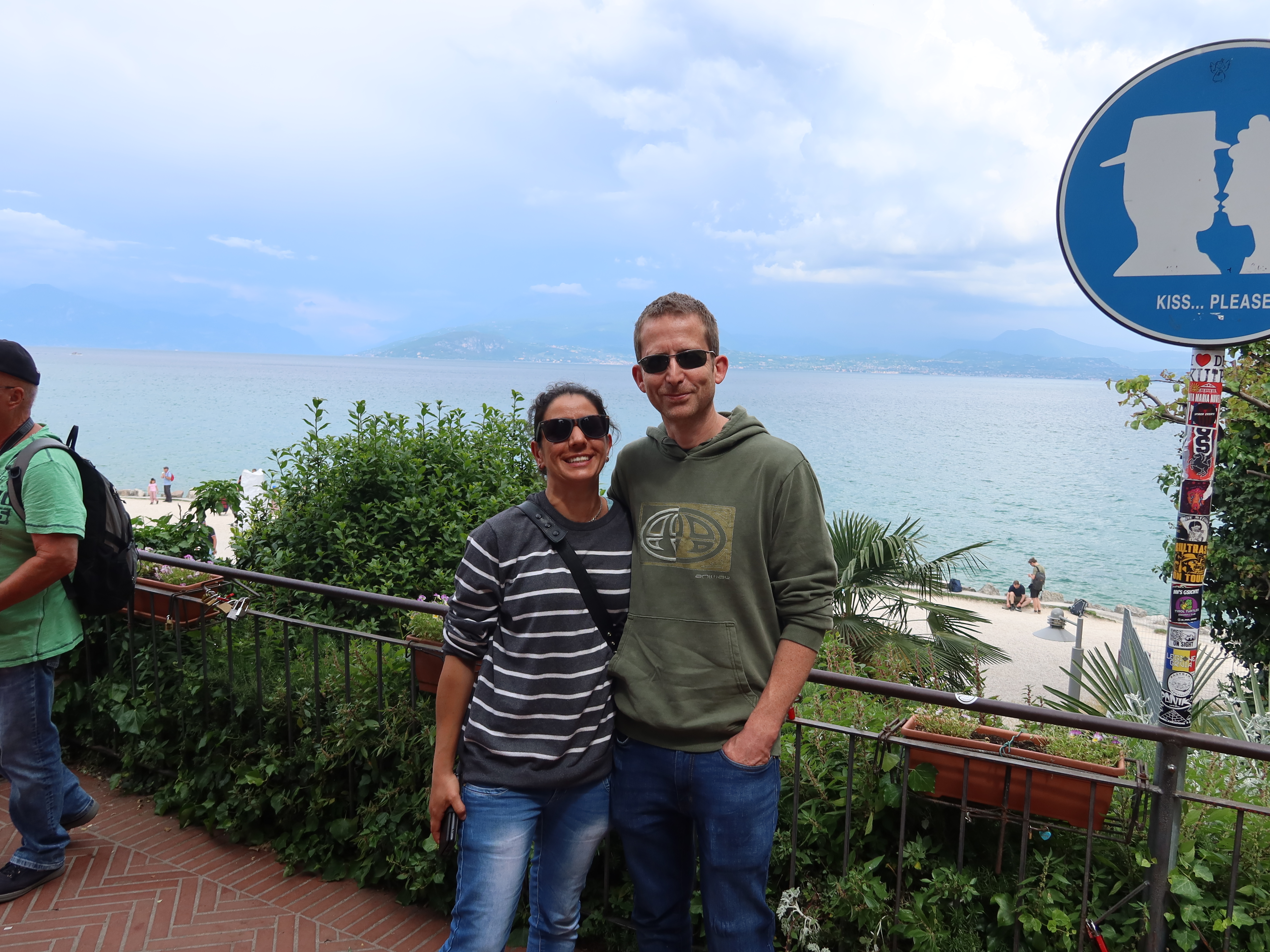 Waterfront Romance Scene Couple smiling by scenic waterfront, blue sign with "KISS... PLEASE" text visible.