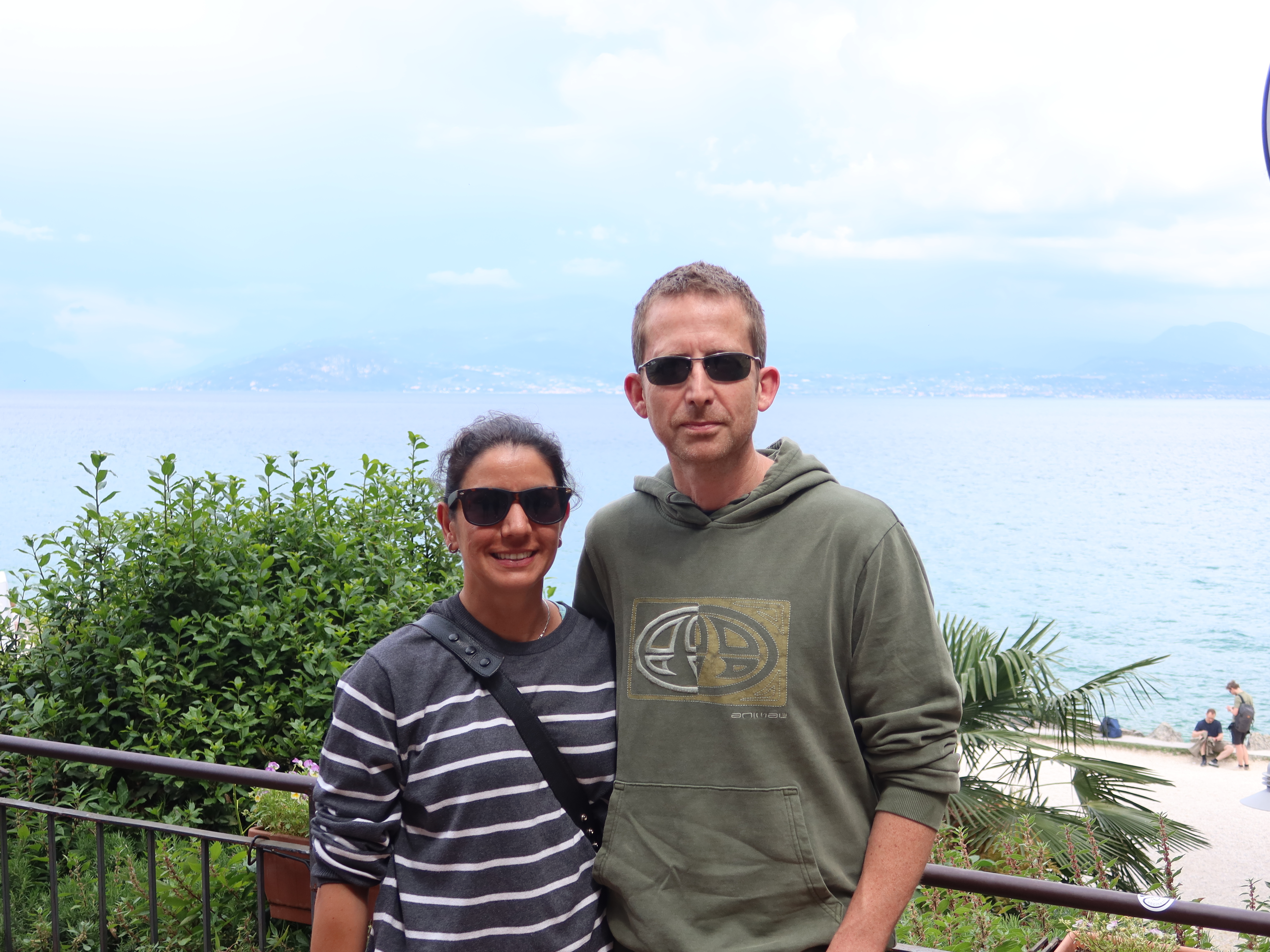 Man and woman in sunglasses outdoors, sea and mountains in the background, green plants nearby.