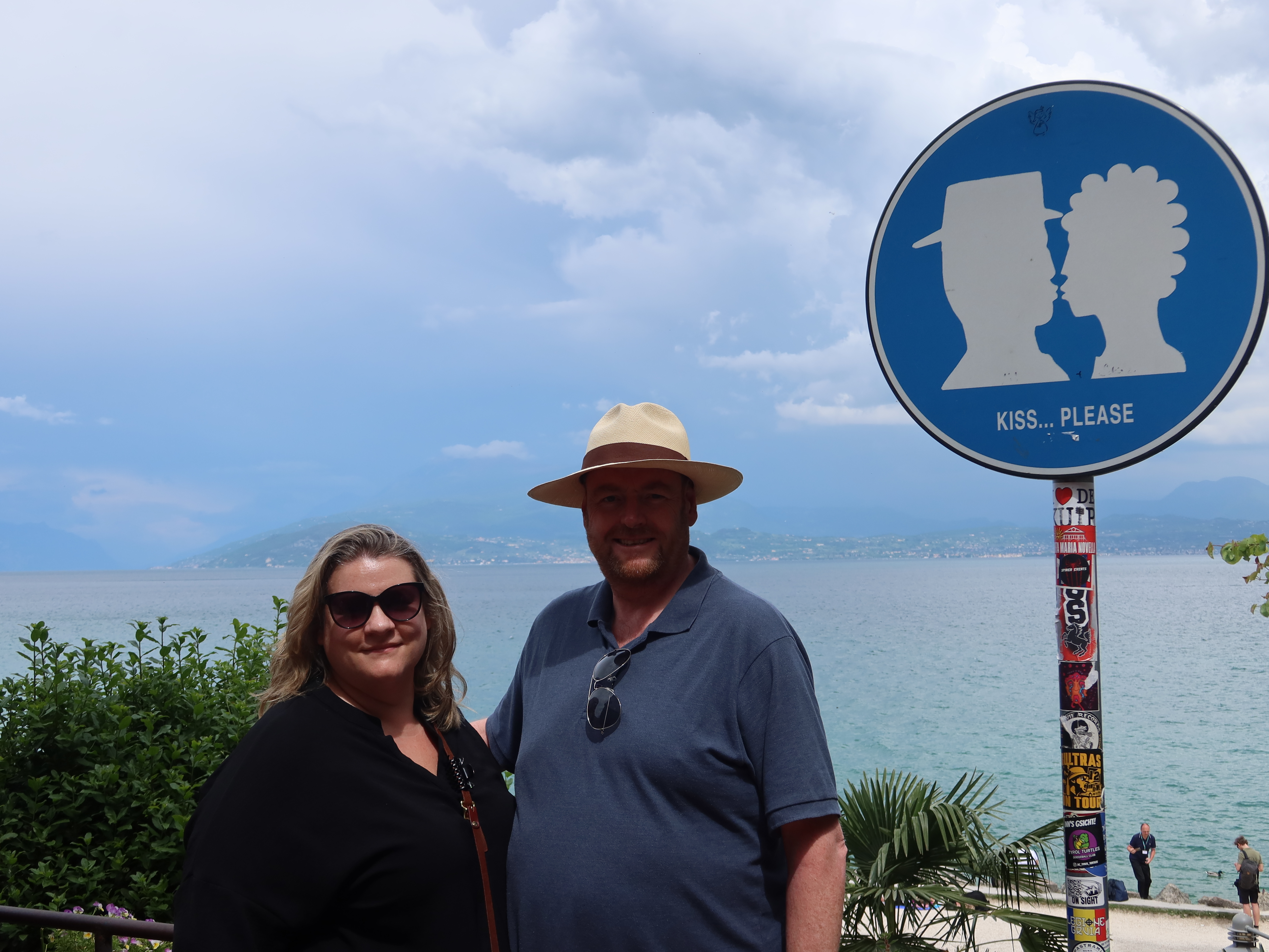 Lakeside Couple with Humorous Sign Couple by lake with humorous "Kiss... Please" sign on sticker-covered pole, cloudy sky backdrop.
