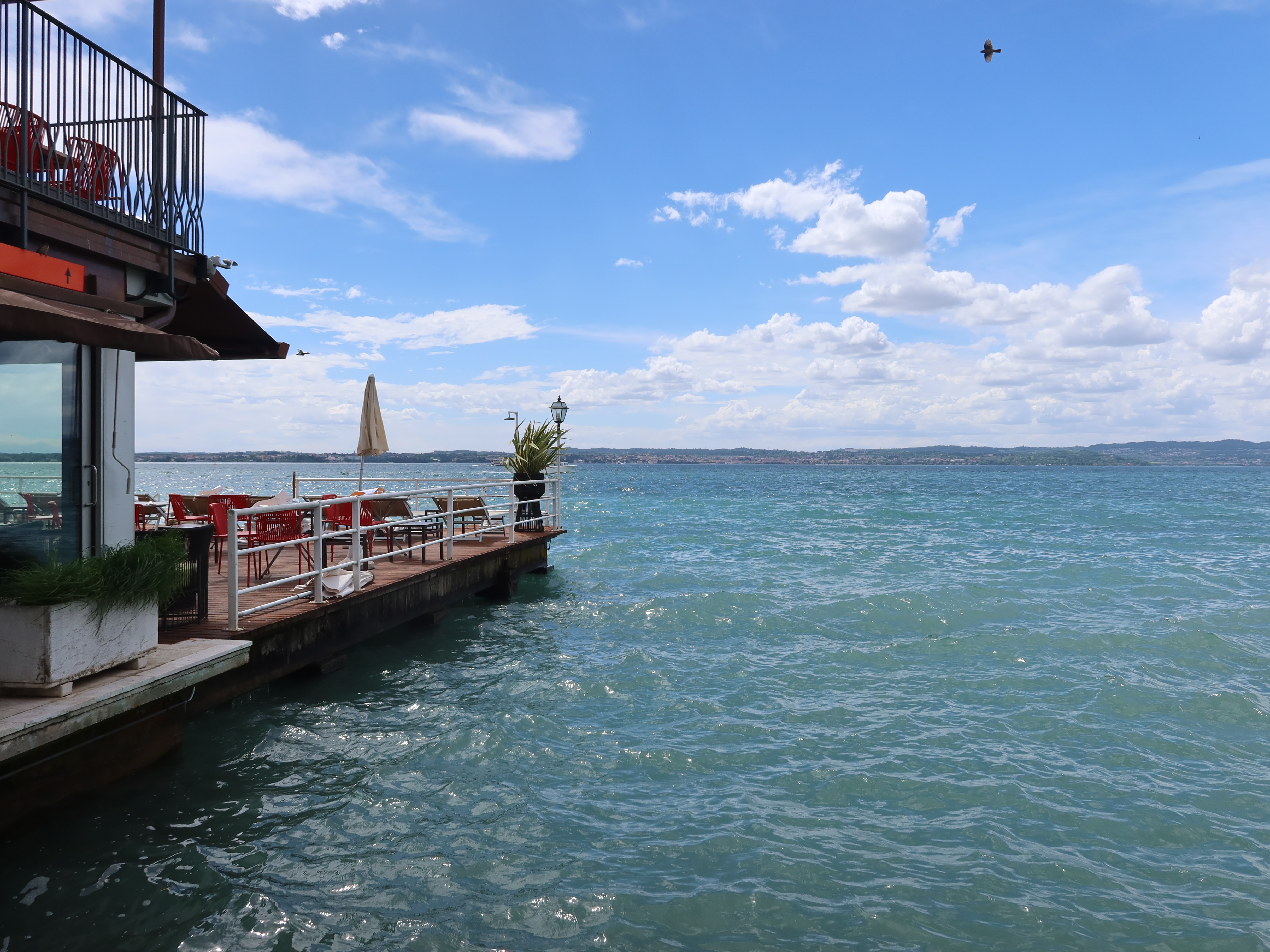 Waterfront deck with tables, chairs, umbrella; blue water, partly cloudy sky, bird, distant shore.