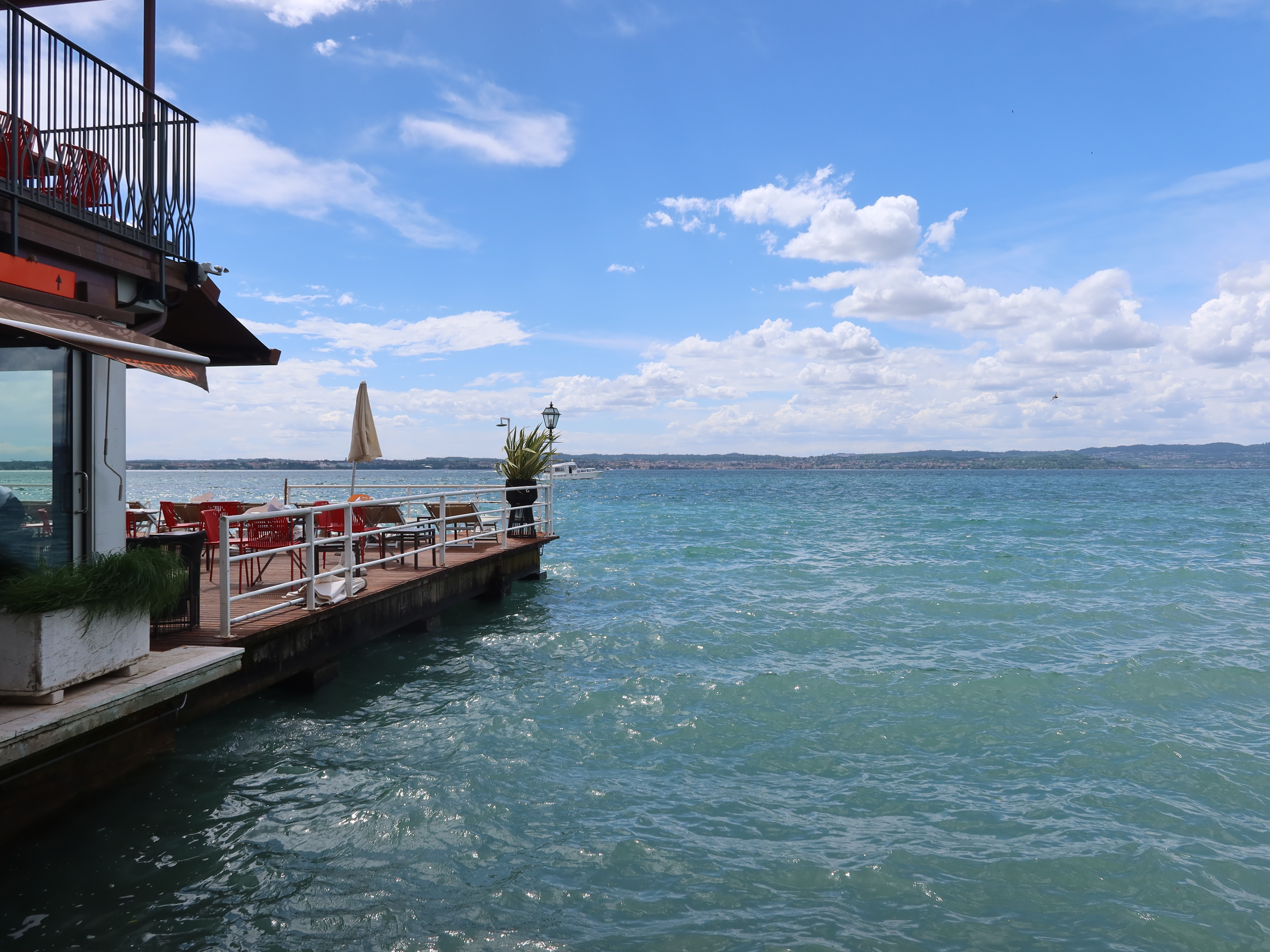 Waterfront deck with red chairs, tables, umbrella, over blue water, hills, and cloudy sky.