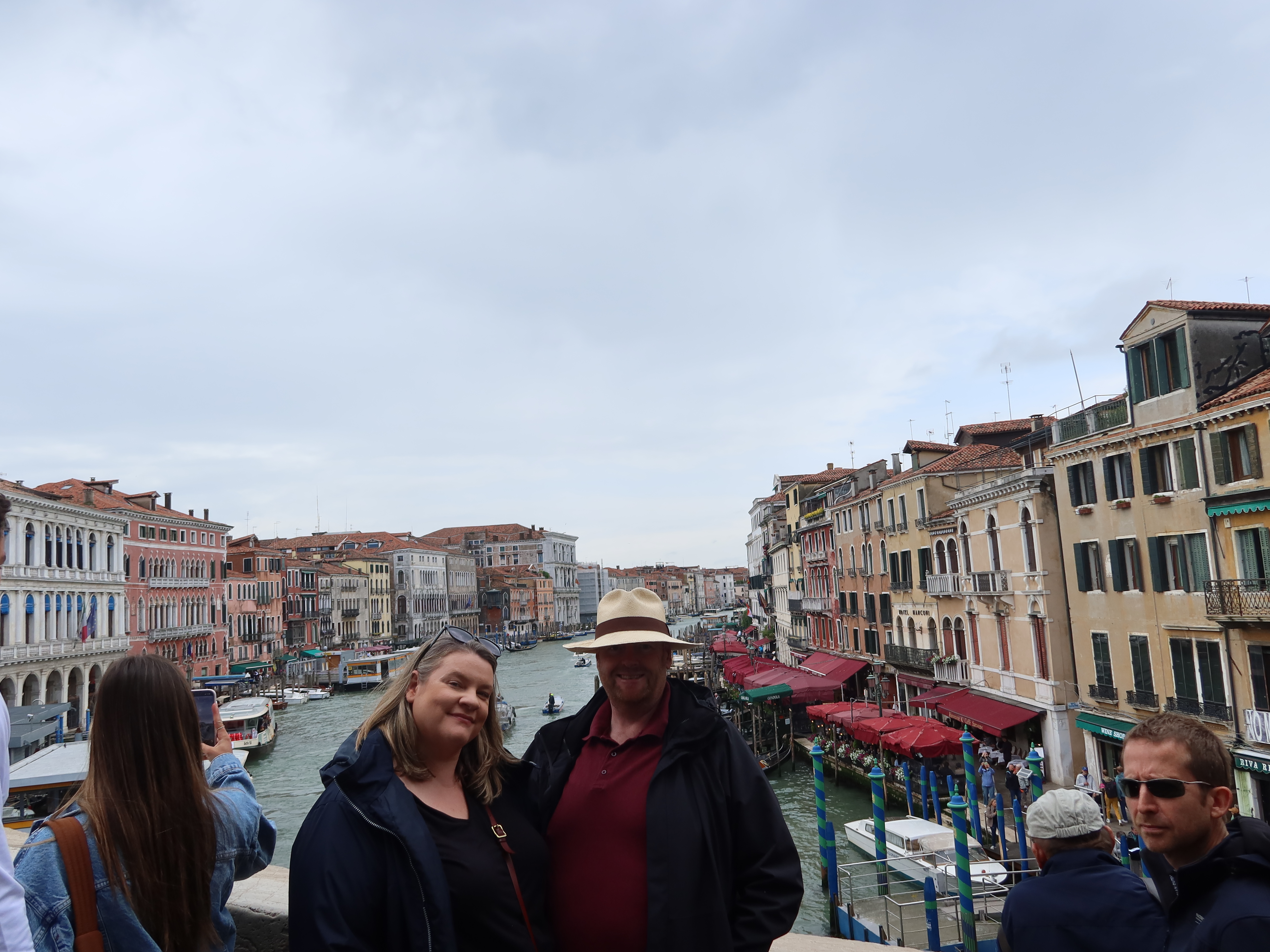 People on a bridge view a canal with colorful buildings and red awnings under an overcast sky.