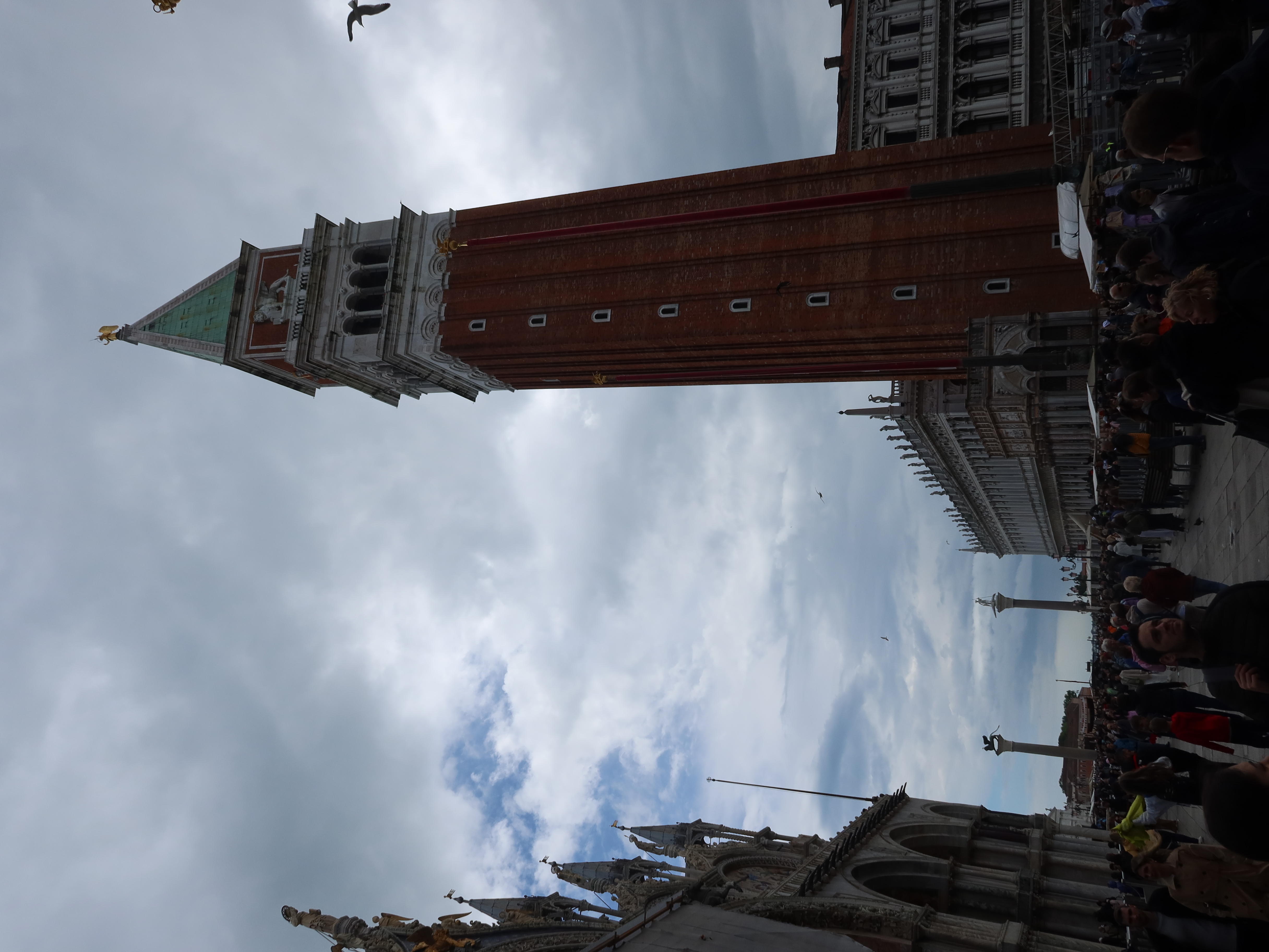 Tall brick bell tower in square with historical buildings, cloudy sky, and a crowd gathering.