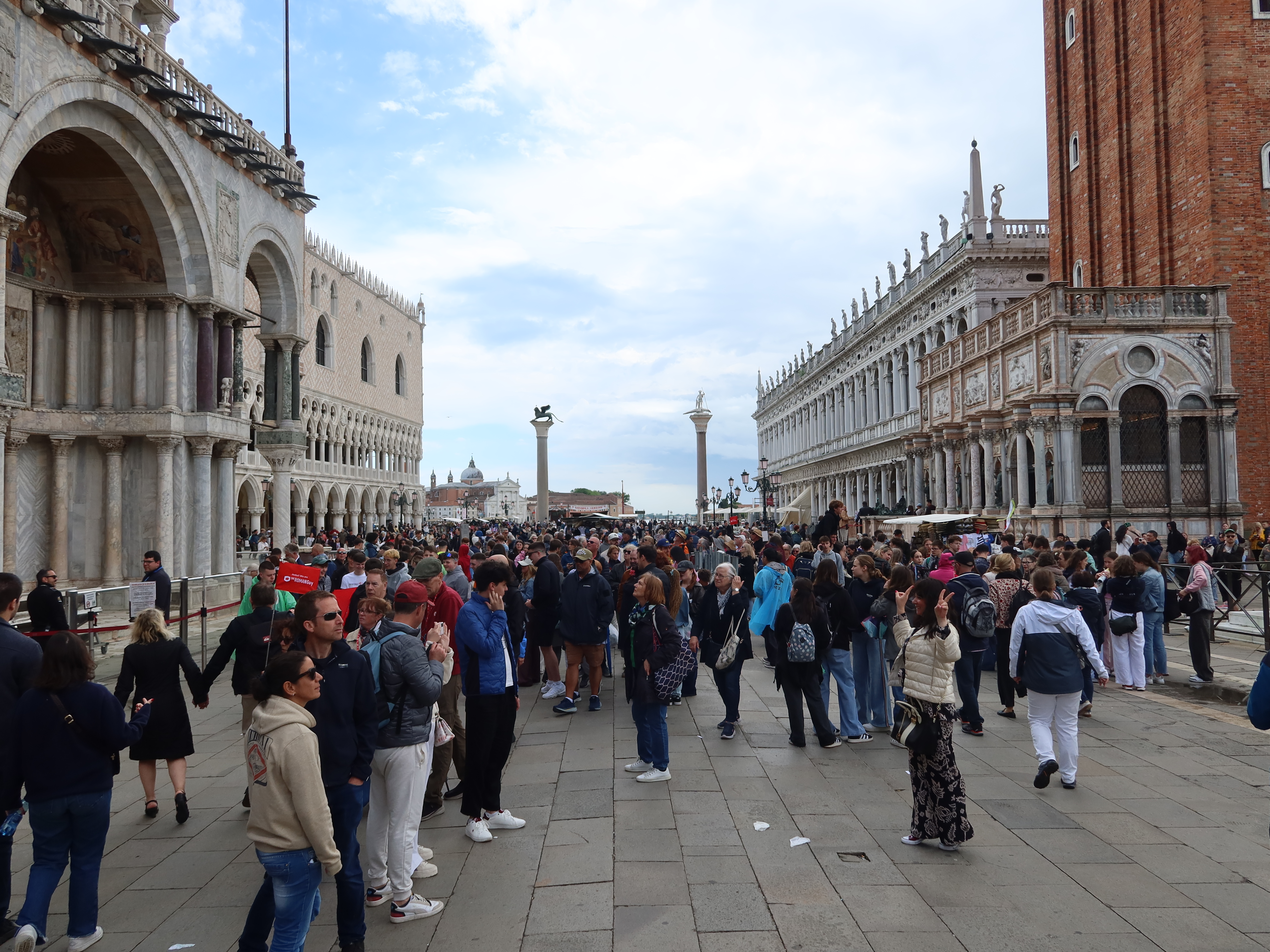 Crowded plaza with historic buildings, archways, columns, and distant tall column on a cloudy day.