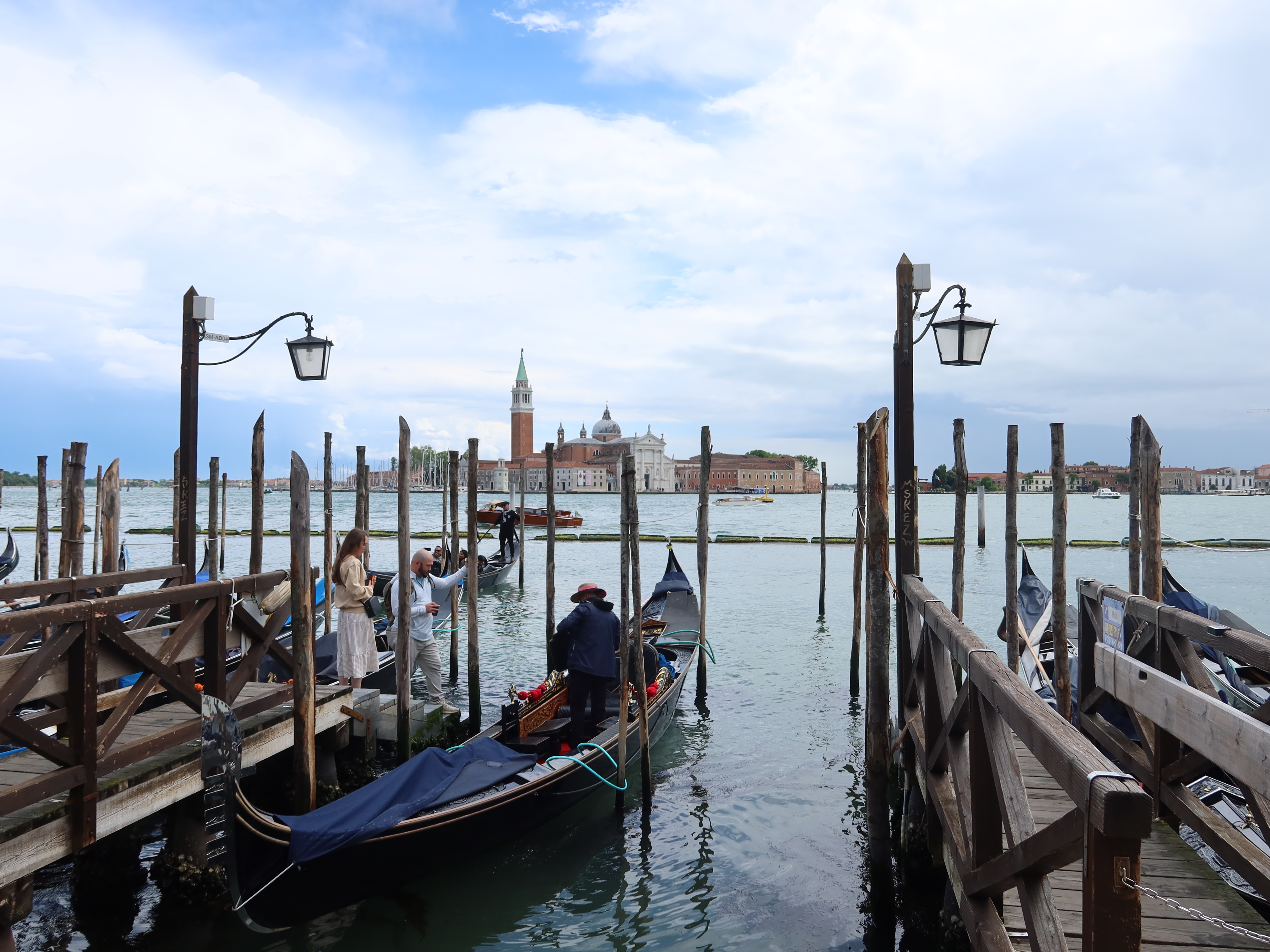 Gondola at a wooden pier with people boarding, historic buildings, and bell tower visible.