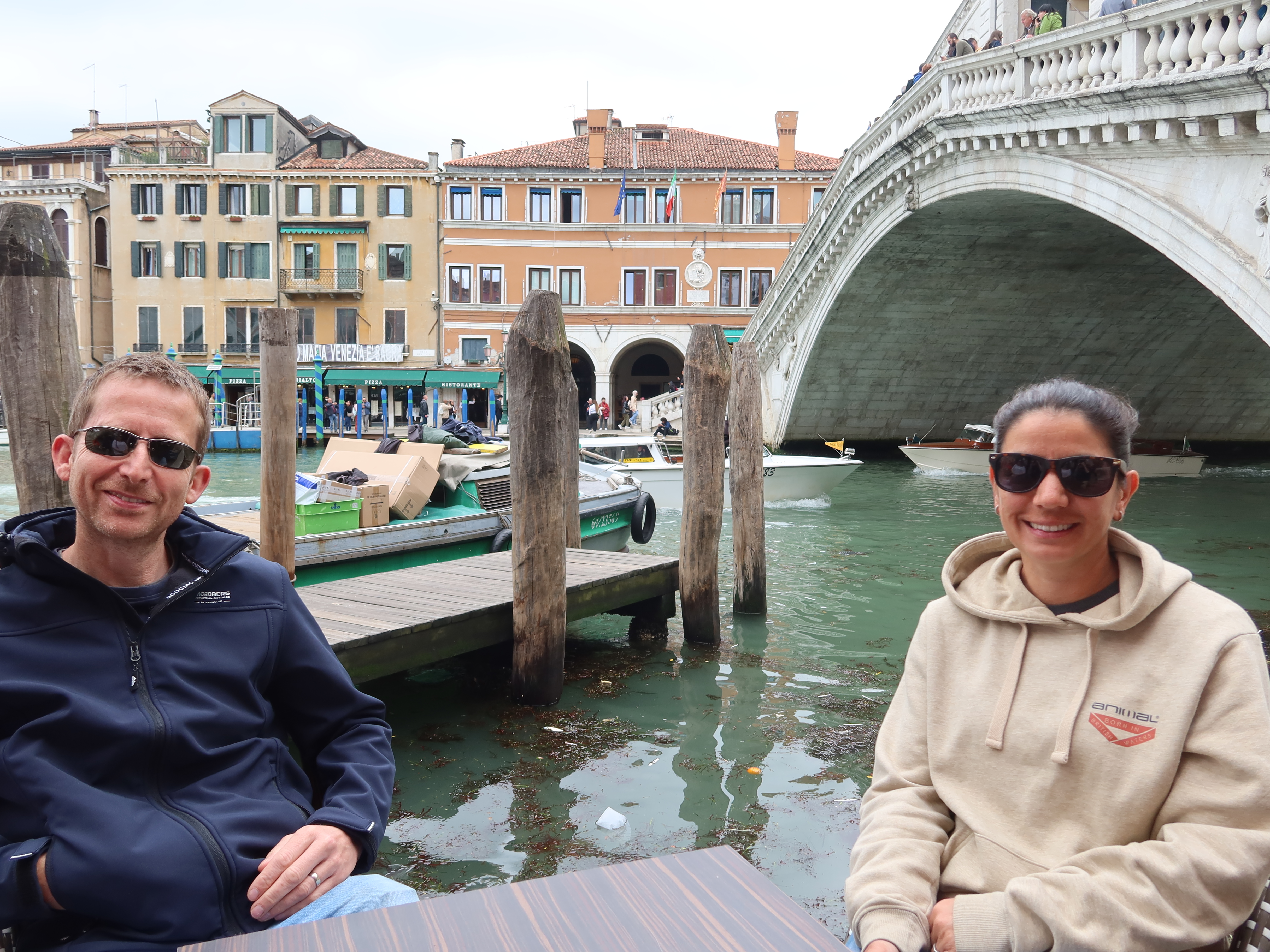 Venice Canal View with Rialto Bridge Man and woman seated near Venice canal with Rialto Bridge and boats in background.
