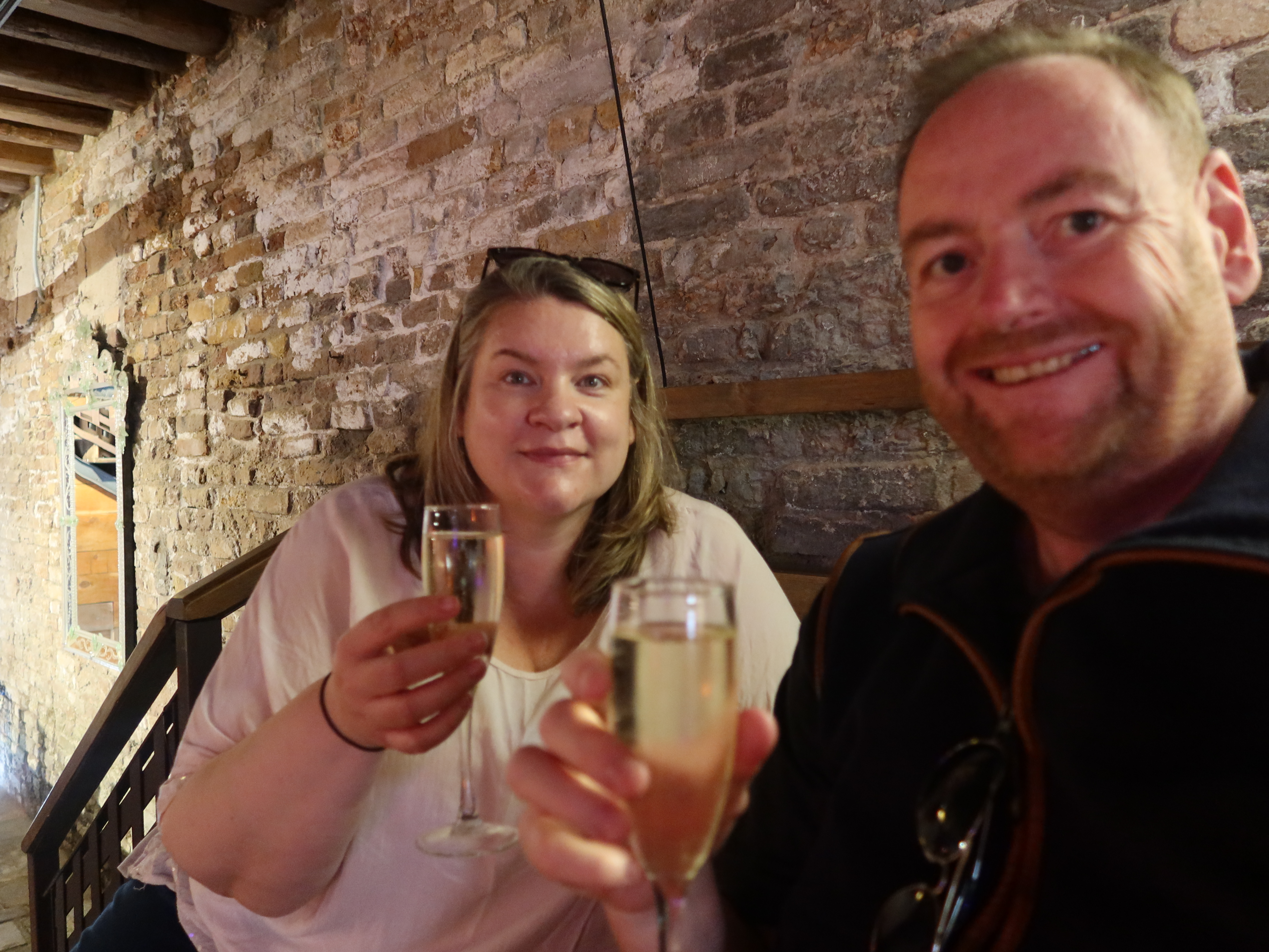 Man and woman smiling, holding champagne glasses in rustic setting with brick walls.