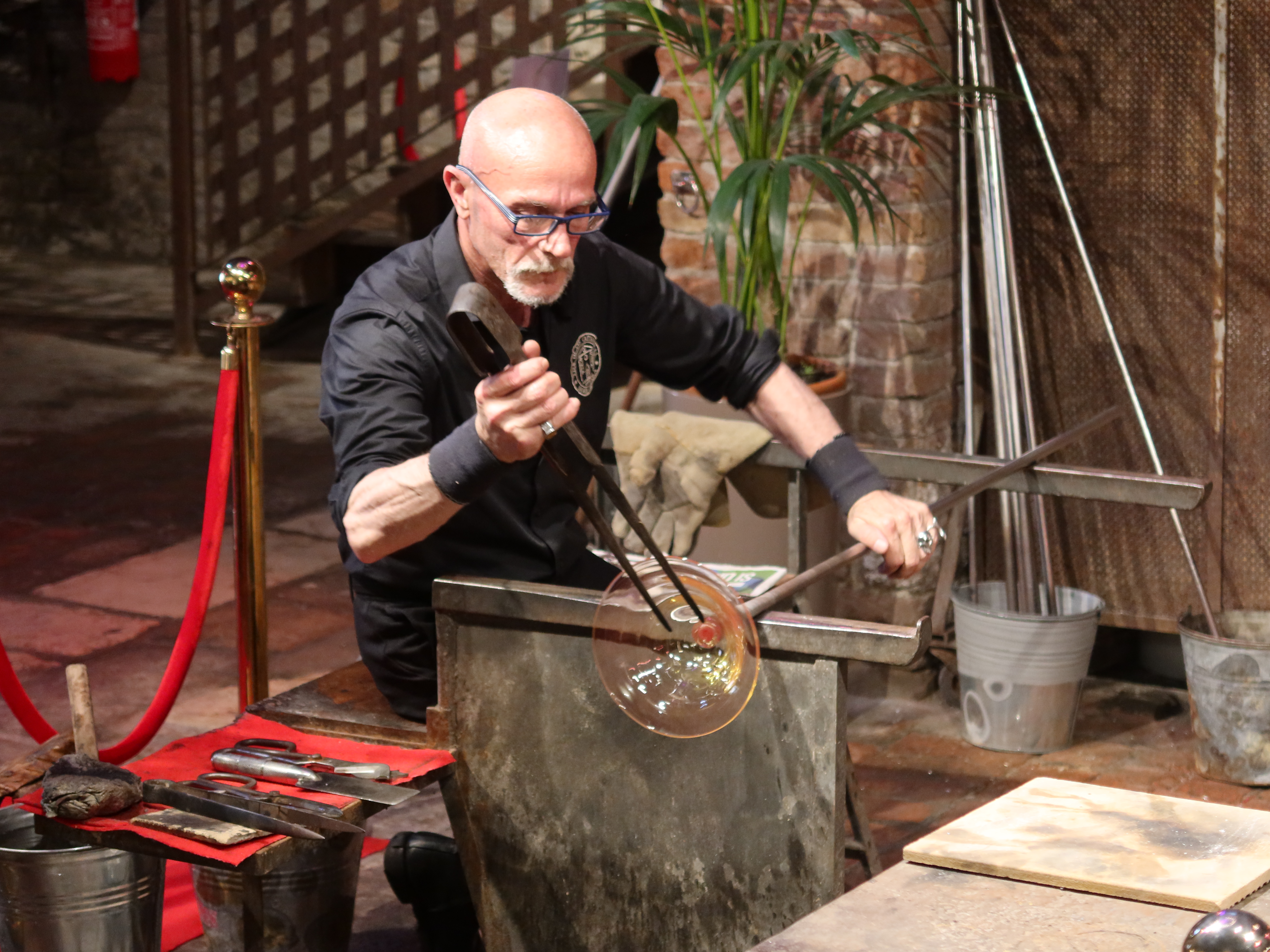 Person glassblowing in workshop with tools, red rope, potted plant, and brick wall visible.