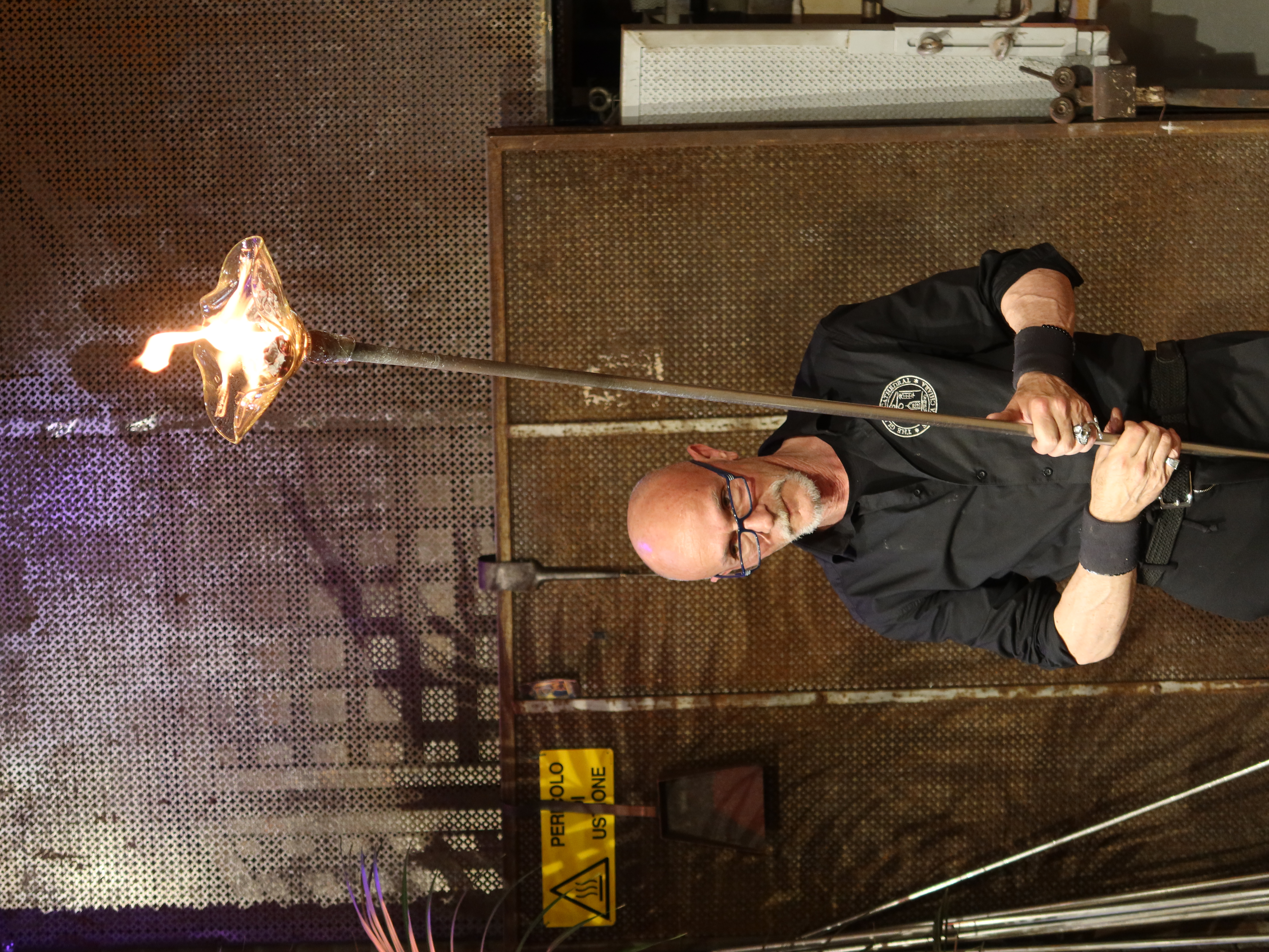 Person with metal rod and flaming glass object against industrial metal wall backdrop.