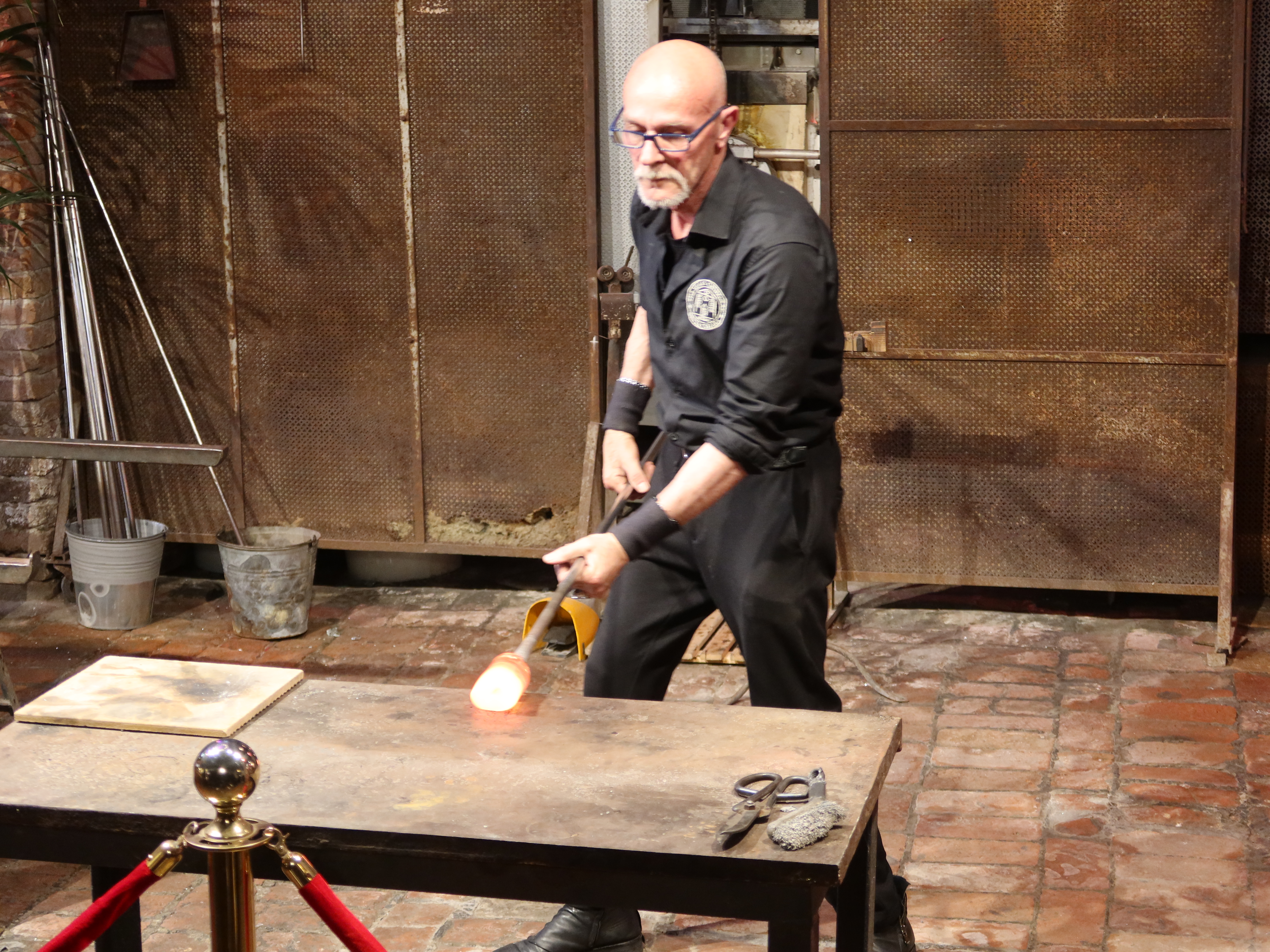 Person glassblowing with molten glass on rod in an industrial setting with tools on table.