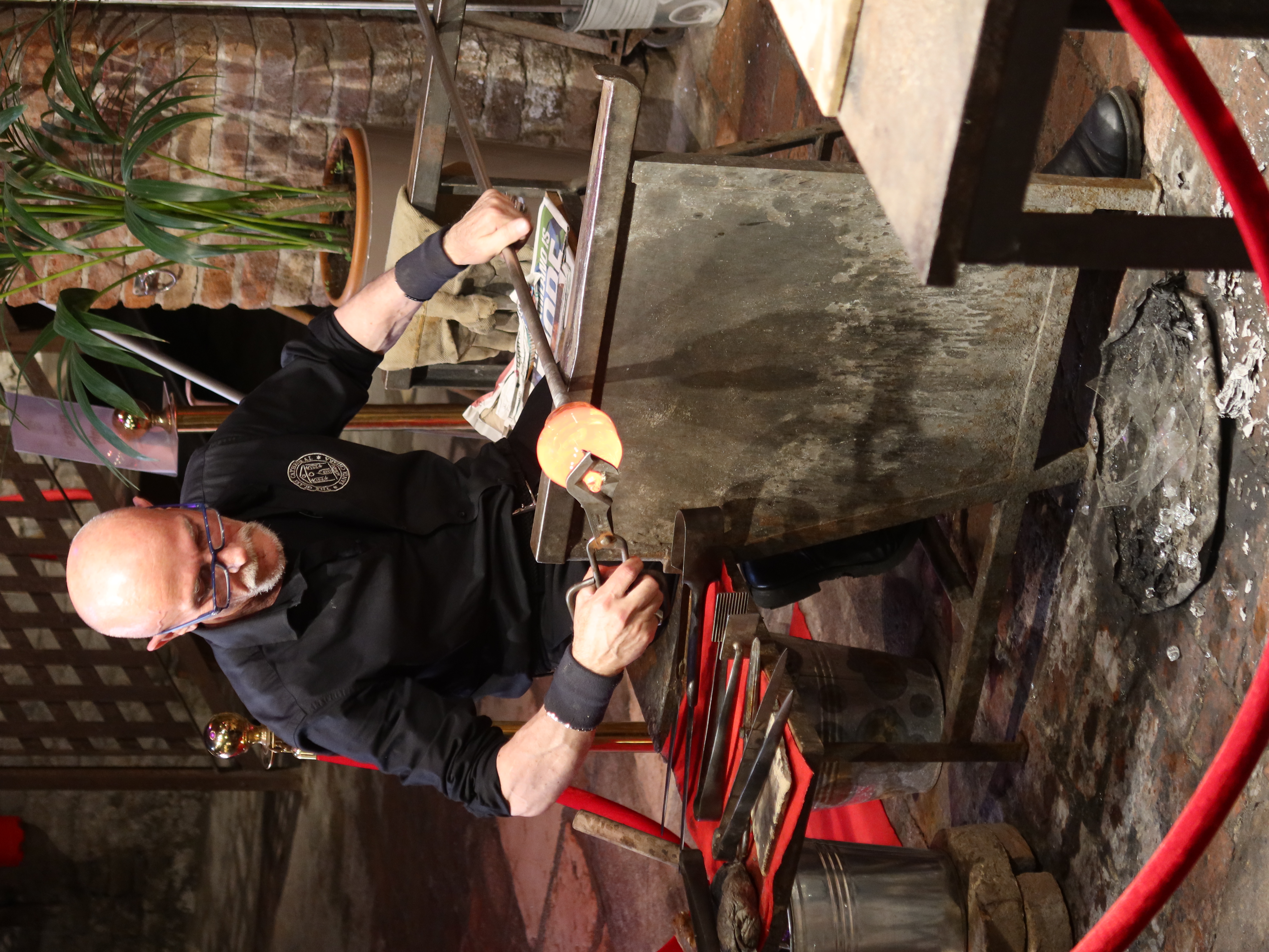Person glassblowing in workshop with tools, protective barrier, and nearby plant.