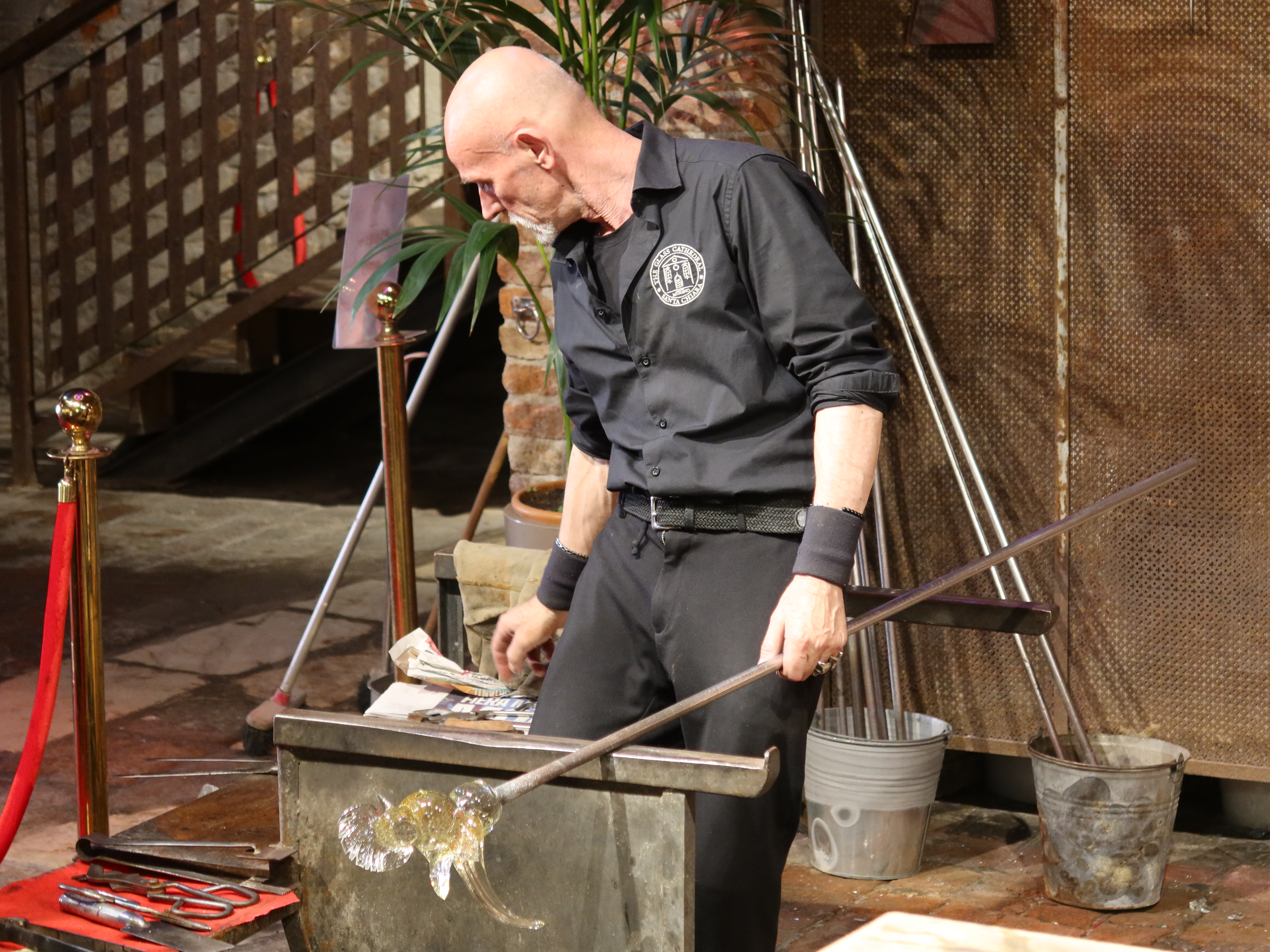 Person in black glassblowing, holding rod with molten glass in workshop with tools and plants.