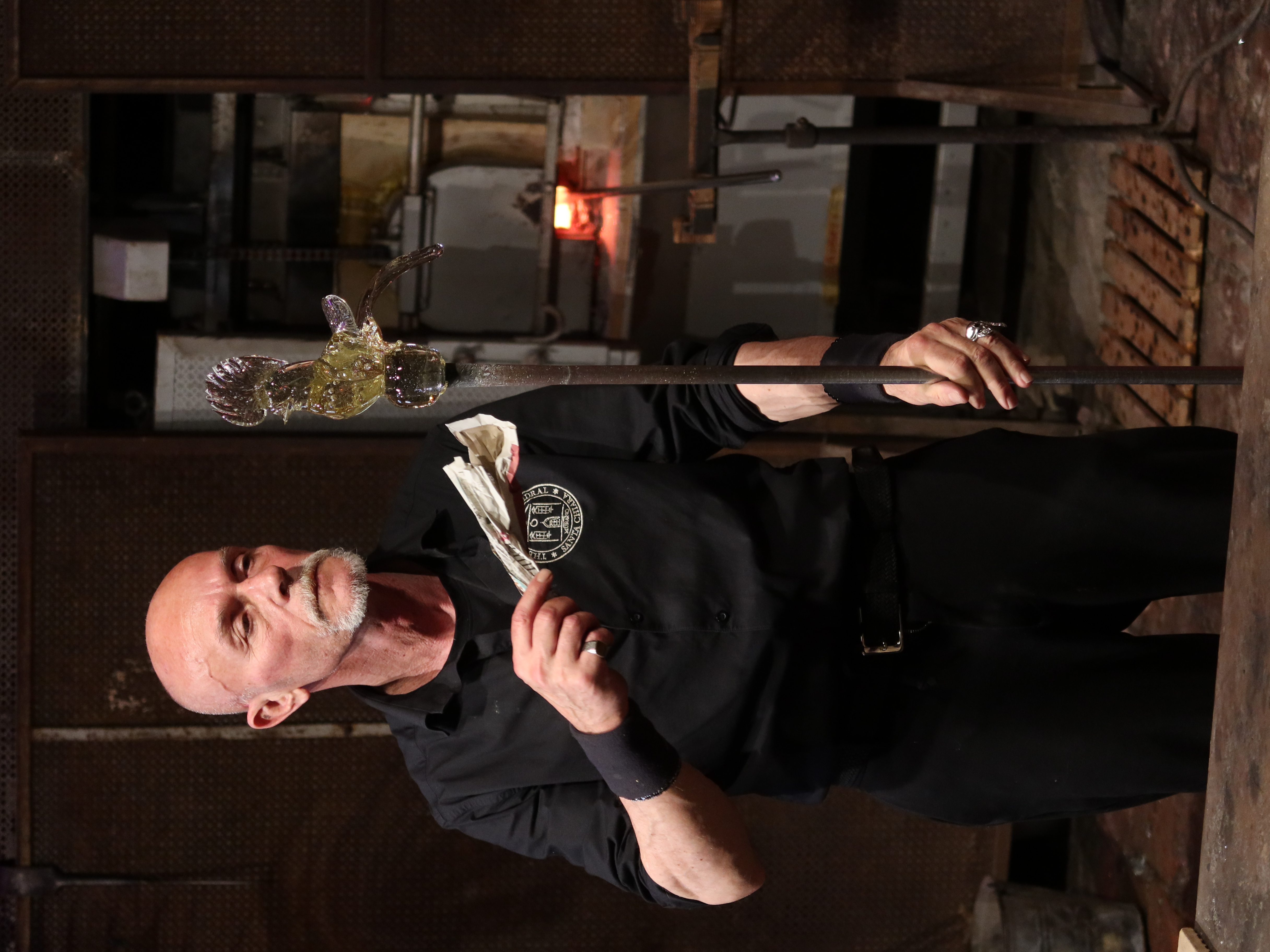 Glass artist shaping bird sculpture with tools in workshop, furnace glowing in background.