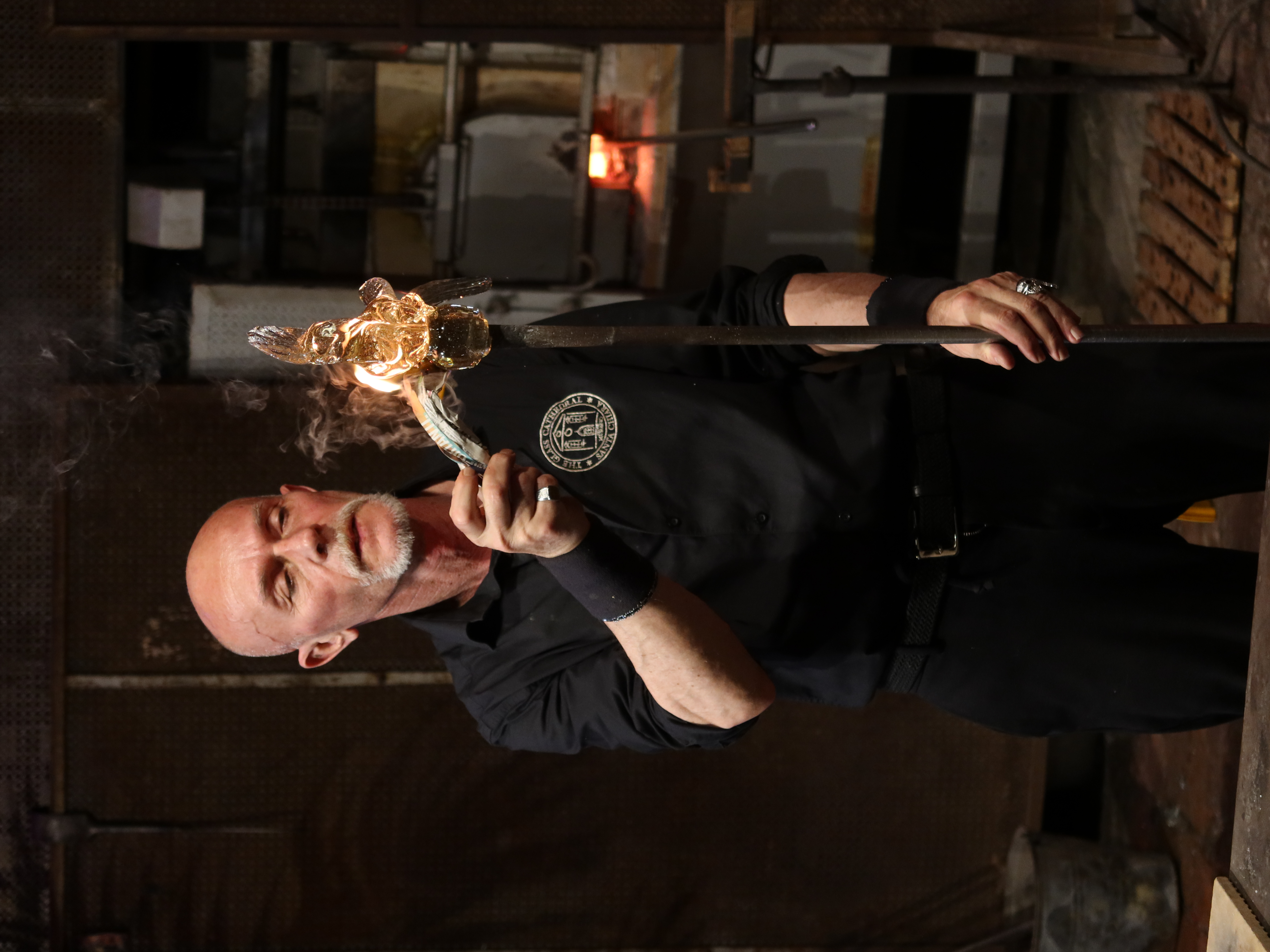 Man shaping glass art with a flame in an industrial setting, wearing a dark shirt and wristbands.