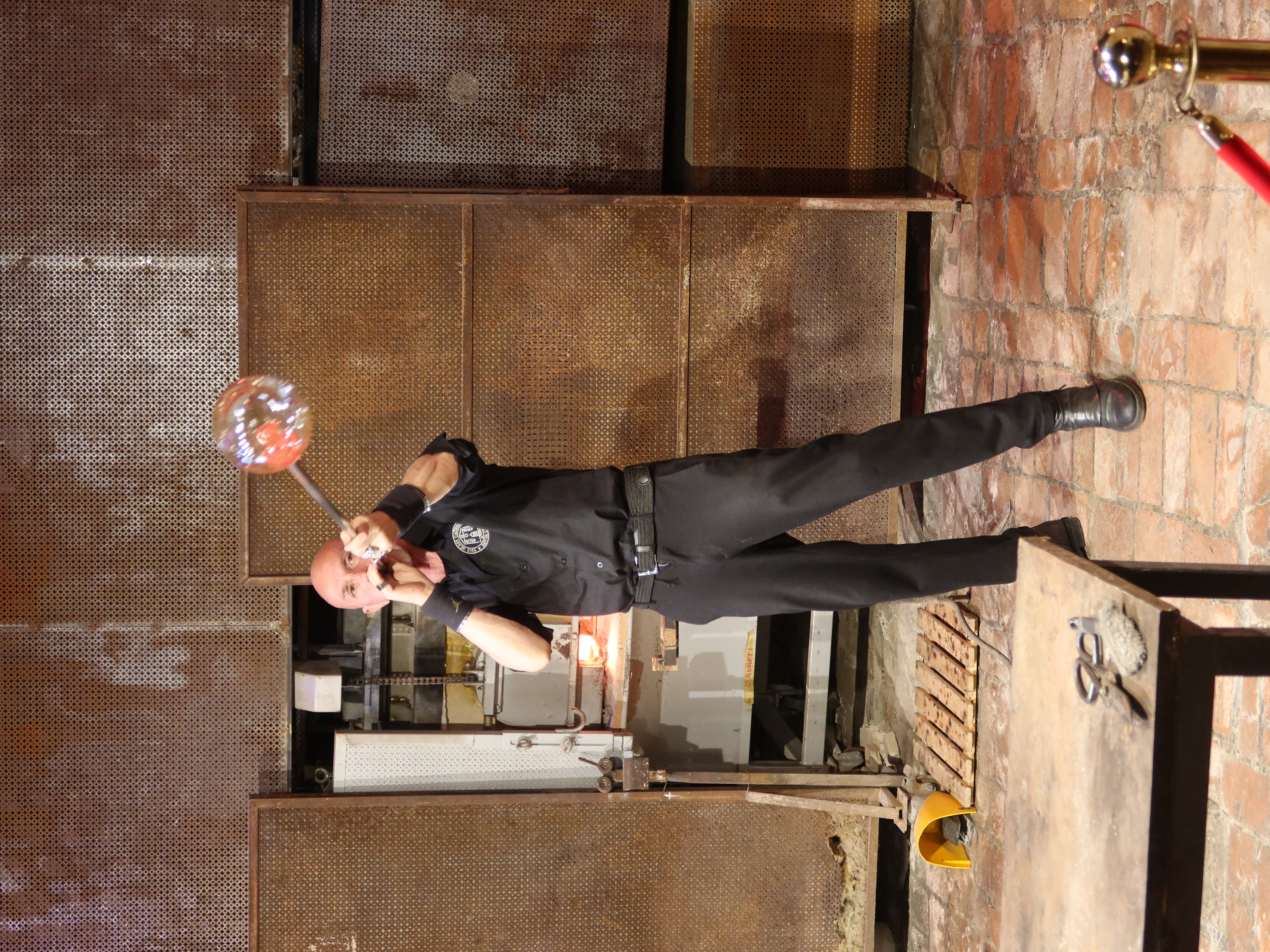 Glassblower shaping molten glass in workshop, tools and furnace visible, wearing black uniform.