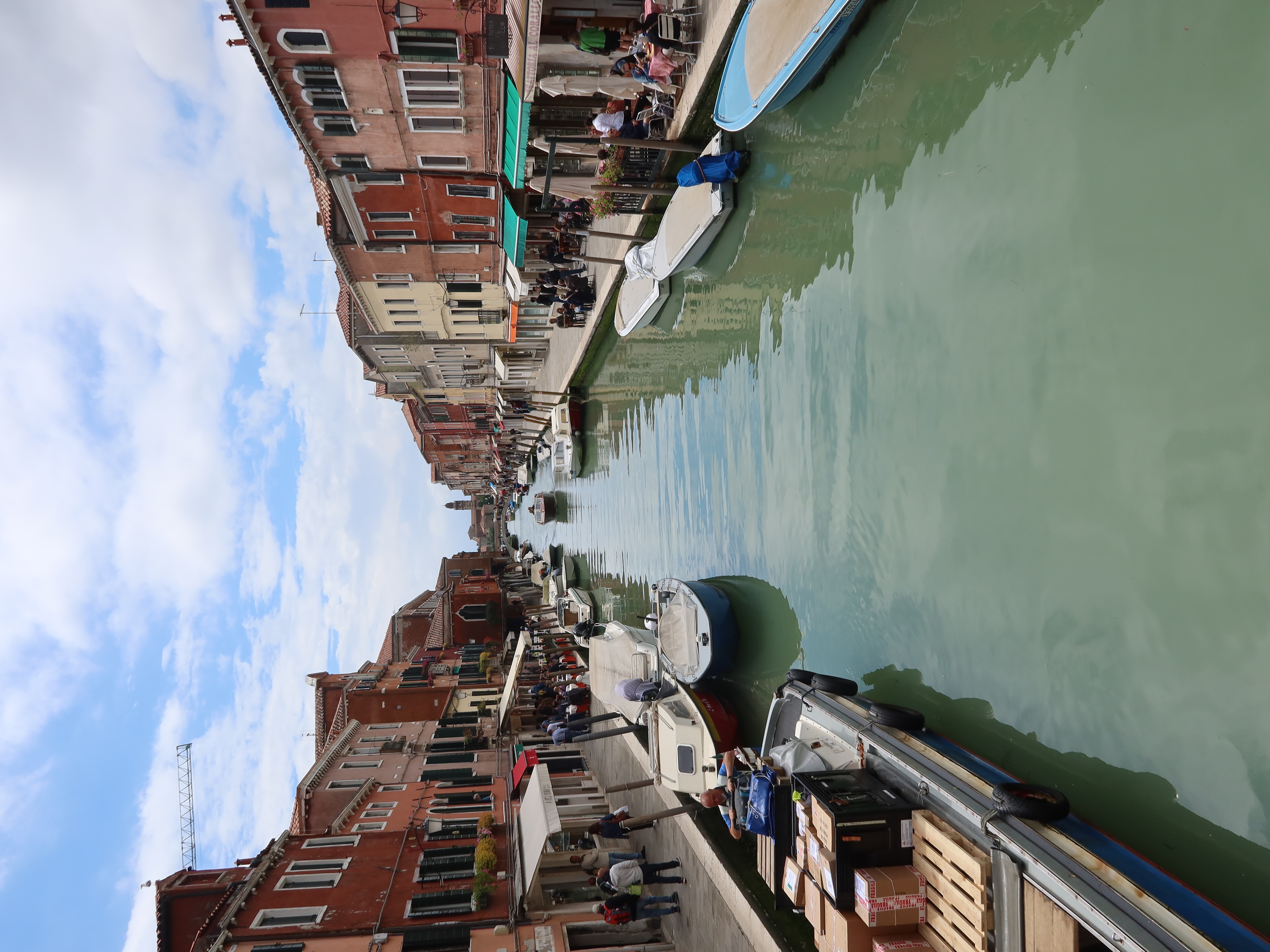 Canal flanked by colorful buildings with docked boats, pedestrians, and outdoor cafes.