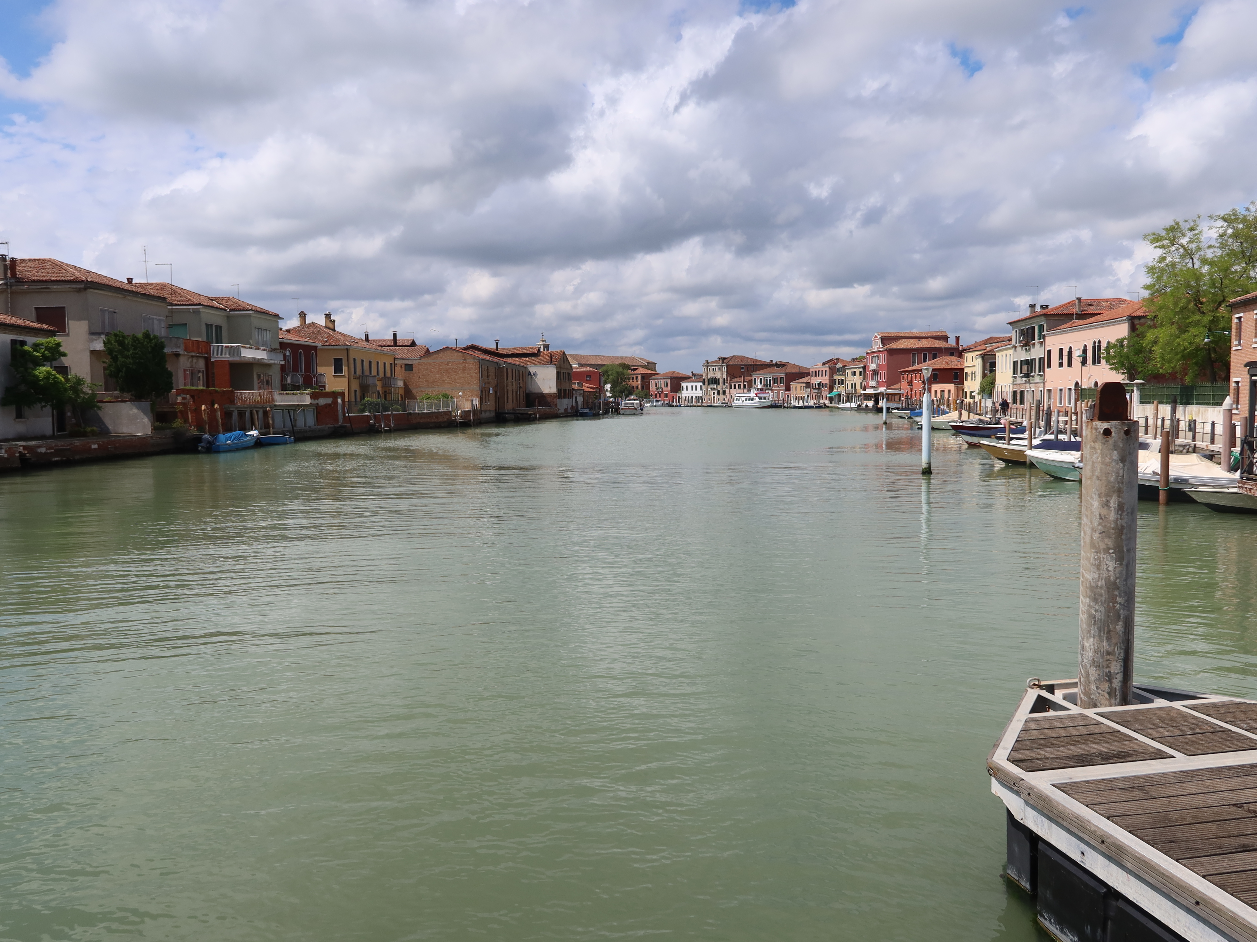 Scenic canal with colorful buildings, moored boats, wooden dock, and partly cloudy sky.