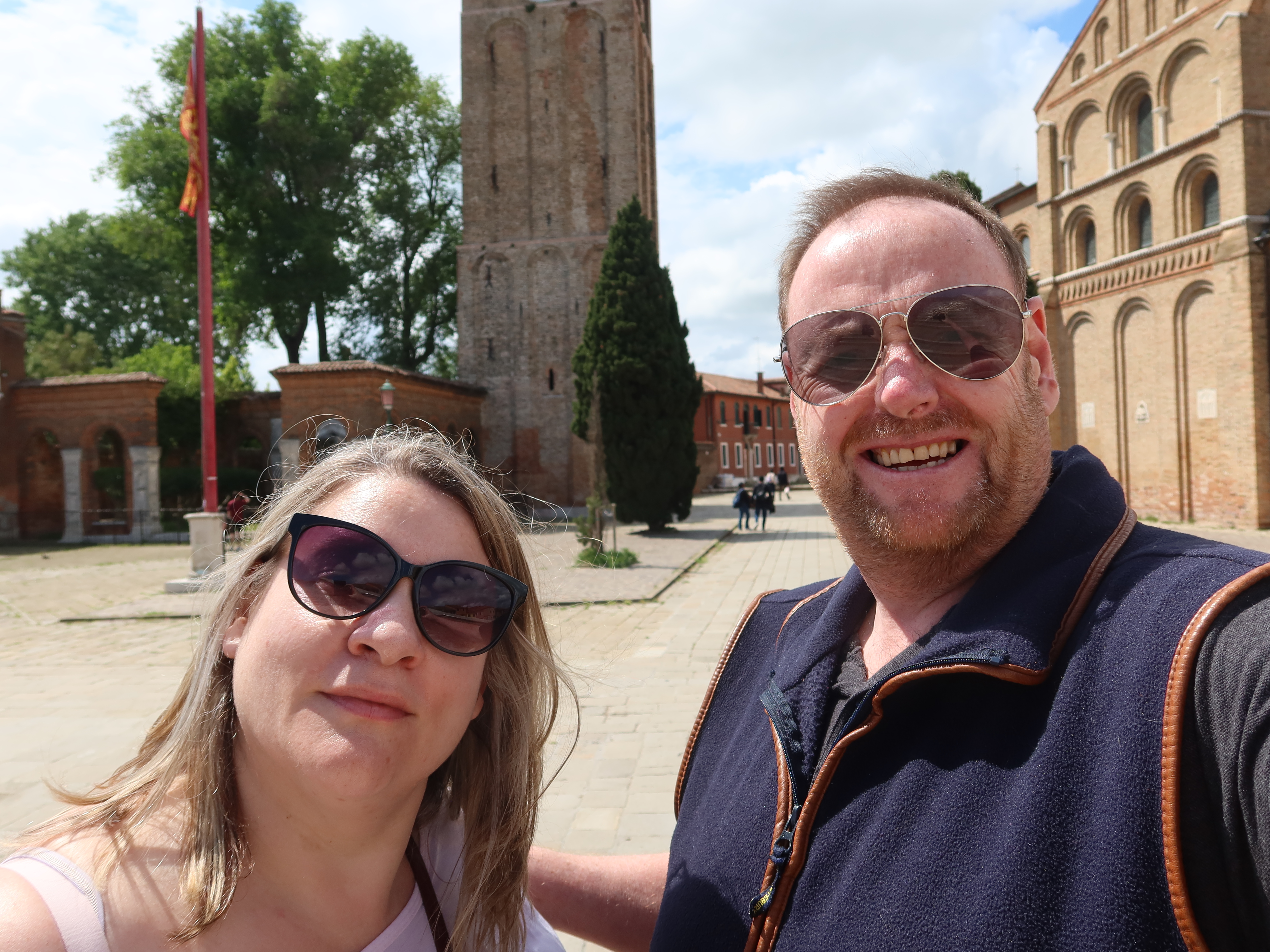 Man and woman wearing sunglasses take a selfie outdoors near a brick tower and arched windows.