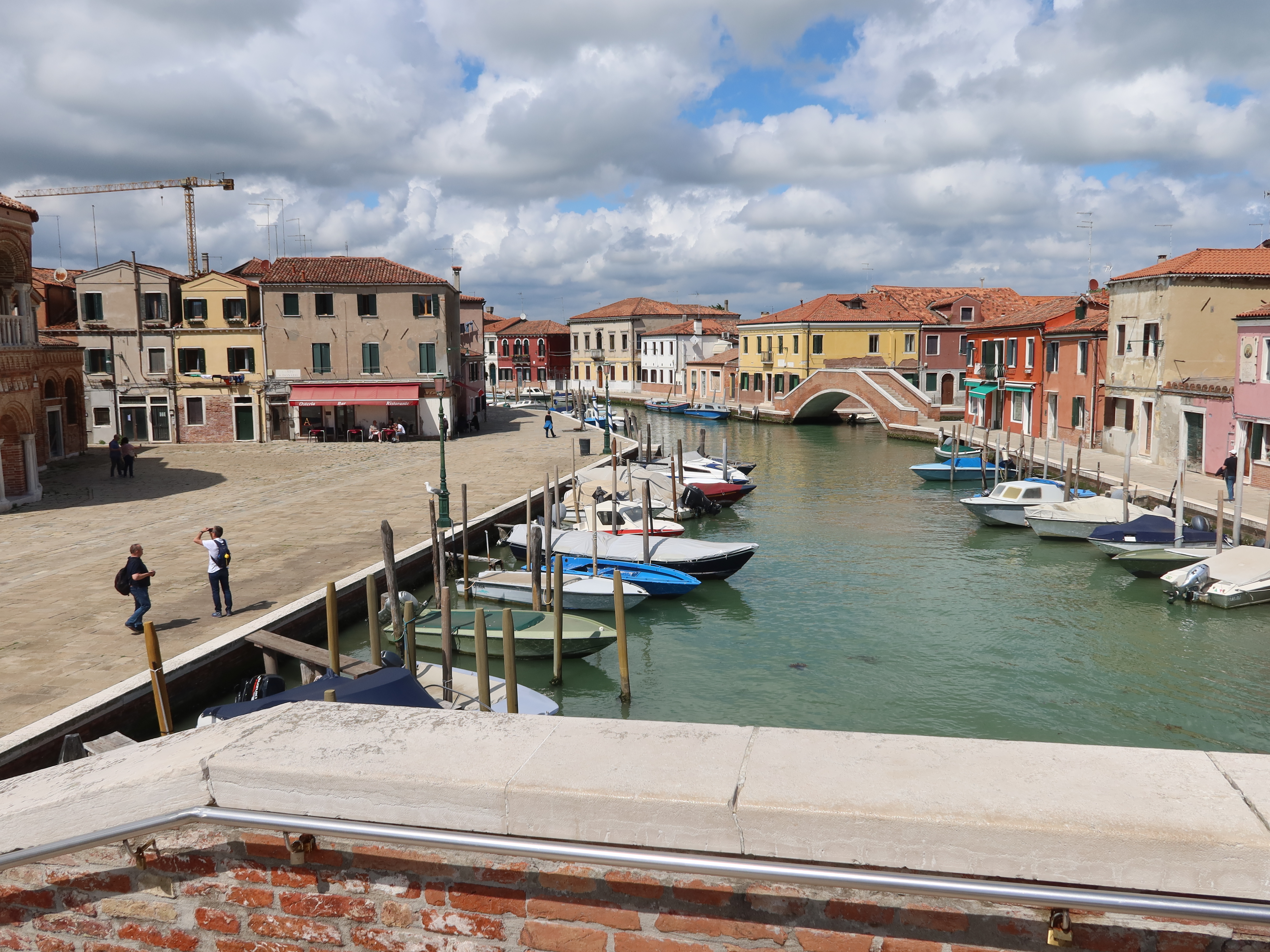 Canal with moored boats, colorful buildings, arched bridge, partly cloudy sky, people nearby.