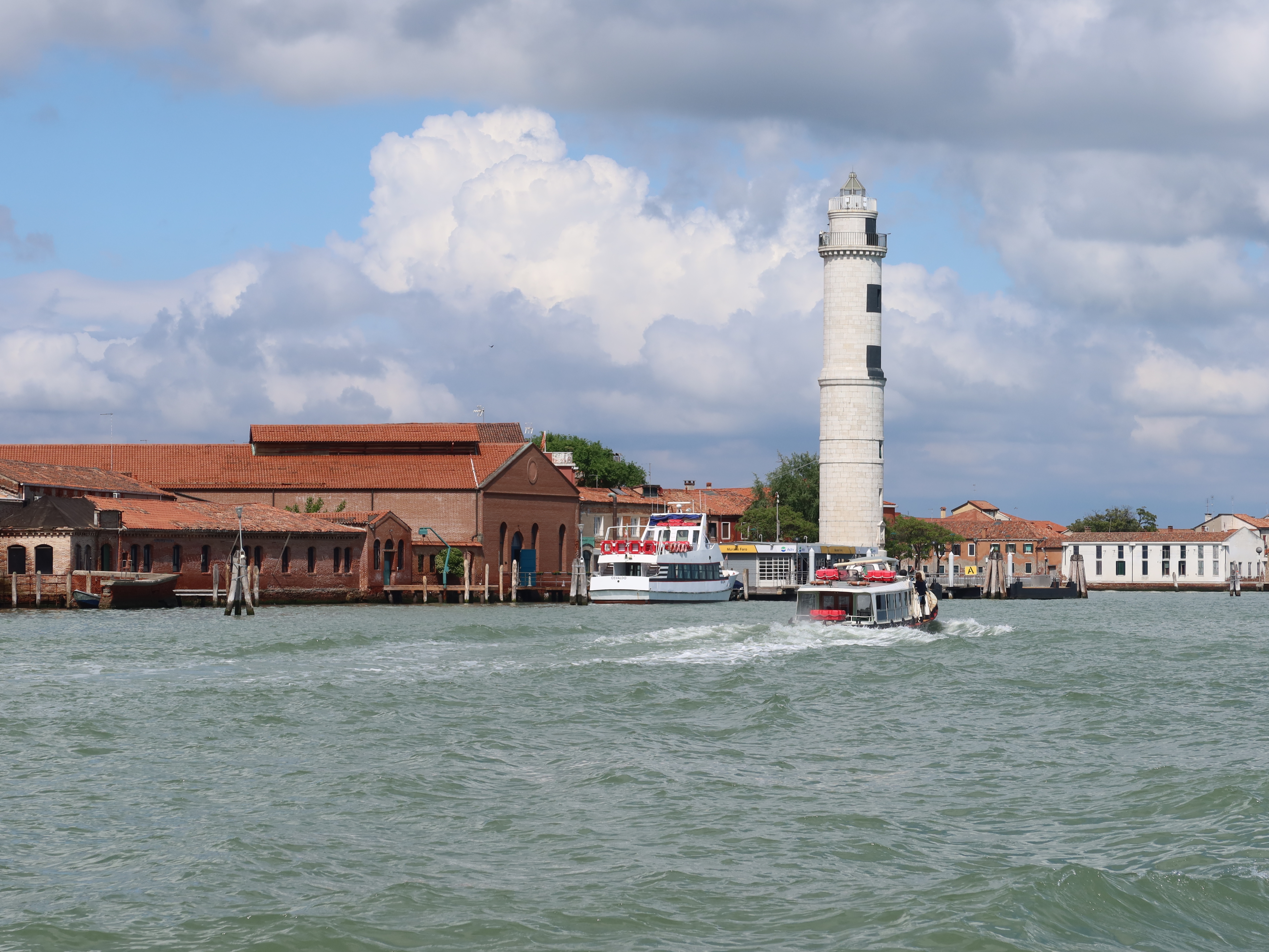 Waterfront scene with a white lighthouse, brick buildings, boats, and partly cloudy sky.