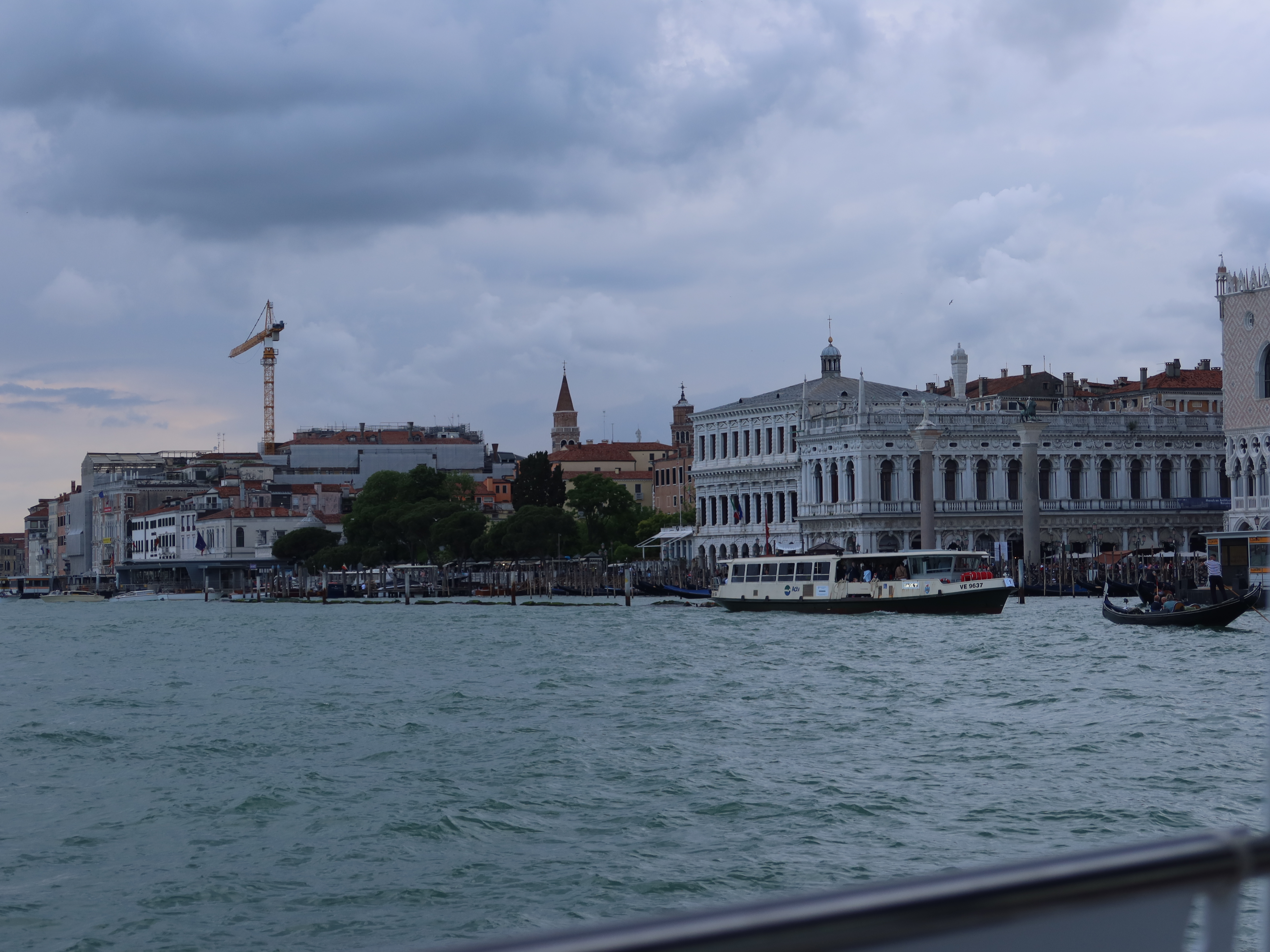Waterfront cityscape with historical buildings, crane, church spire, and boats under cloudy sky.
