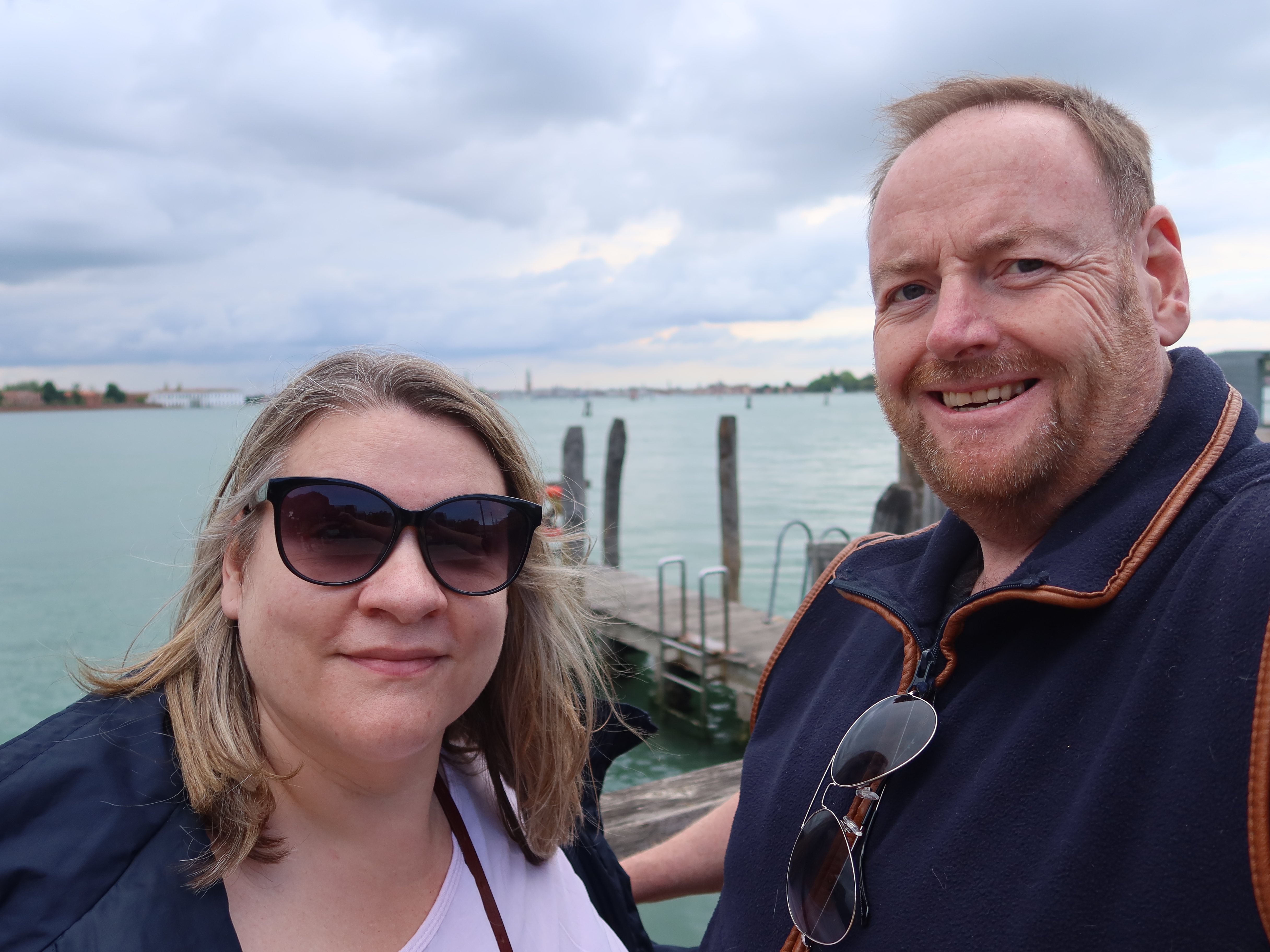 Man and woman taking selfie by waterfront, sunglasses, jacket, cloudy sky, distant shoreline.