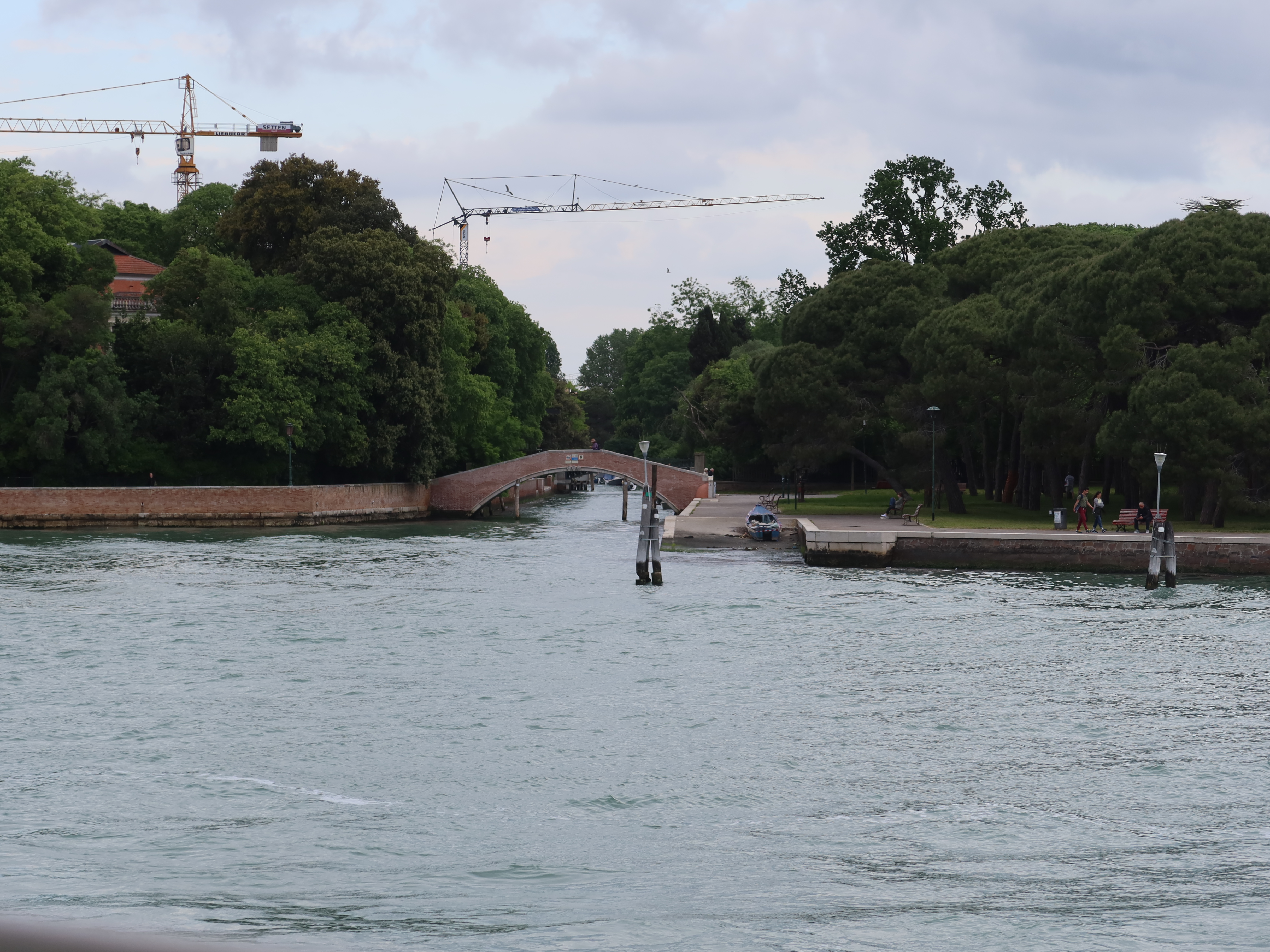 Waterway with arched bridge, greenery, cranes, and people near water under overcast sky.